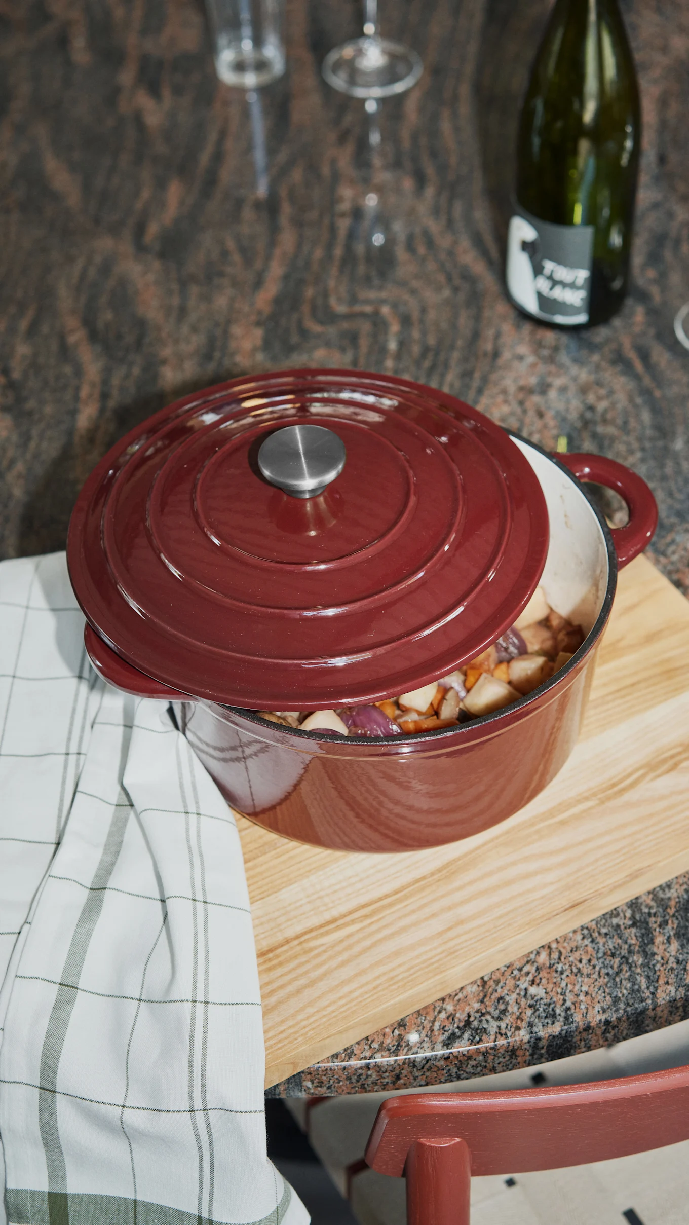 Maroon cast iron pot with lid slightly ajar, revealing chopped vegetables on a wooden board.
