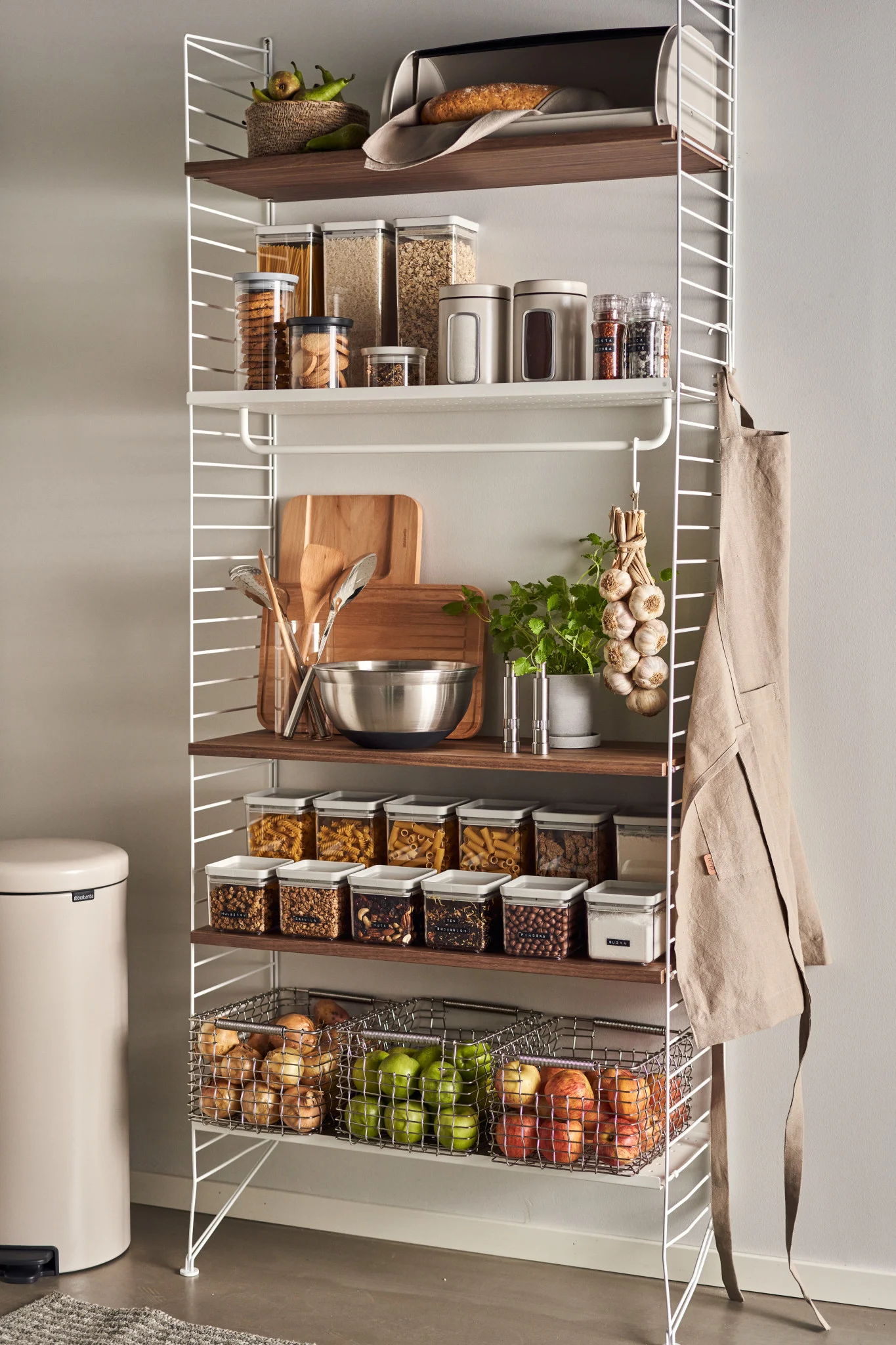 A white shelving unit with wooden shelves holds organized pantry items like dry goods in containers, fruits, vegetables, and kitchen tools.