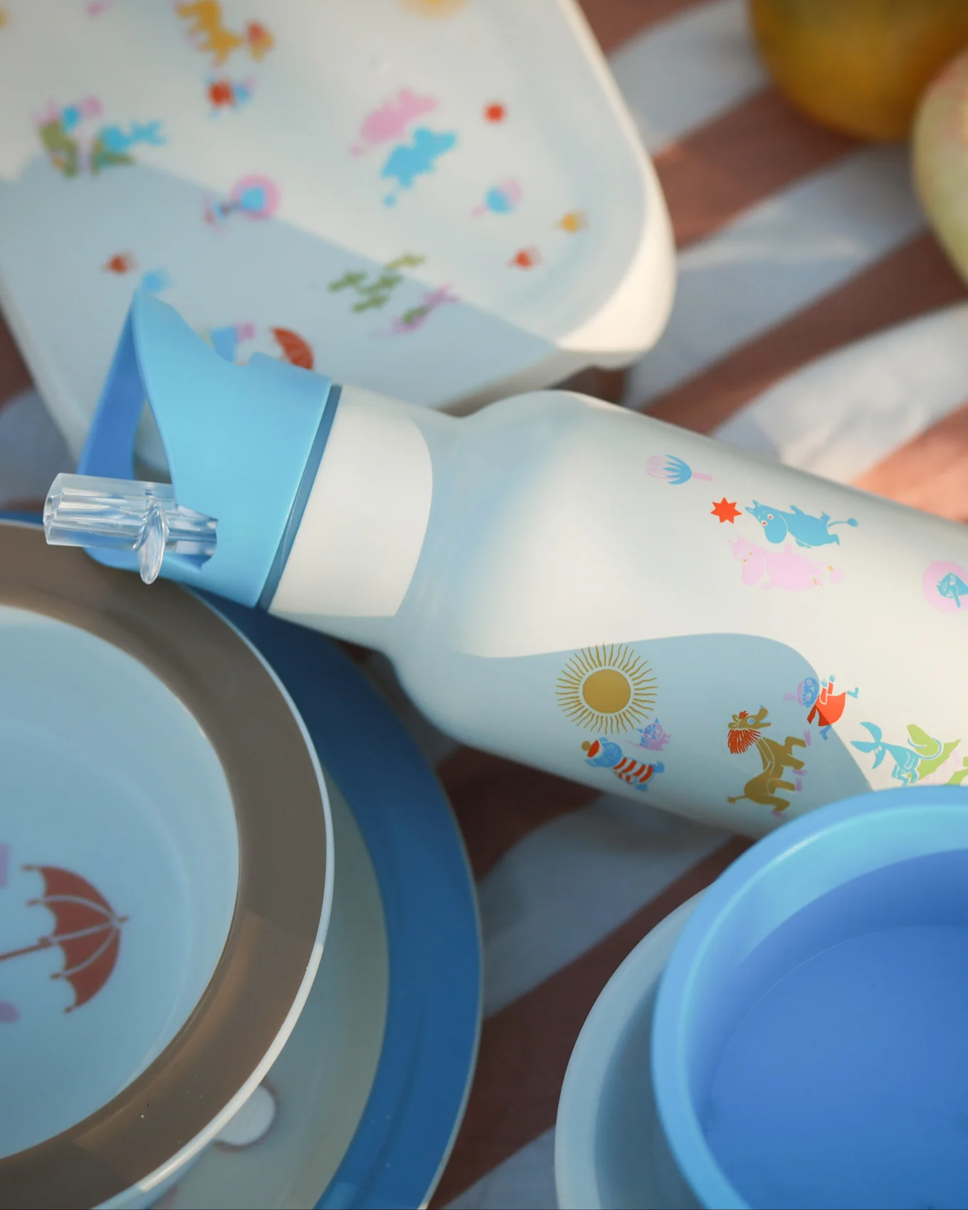 White water bottle with blue straw, colorful cartoon characters, and sun, surrounded by matching picnic plates on a striped blanket.