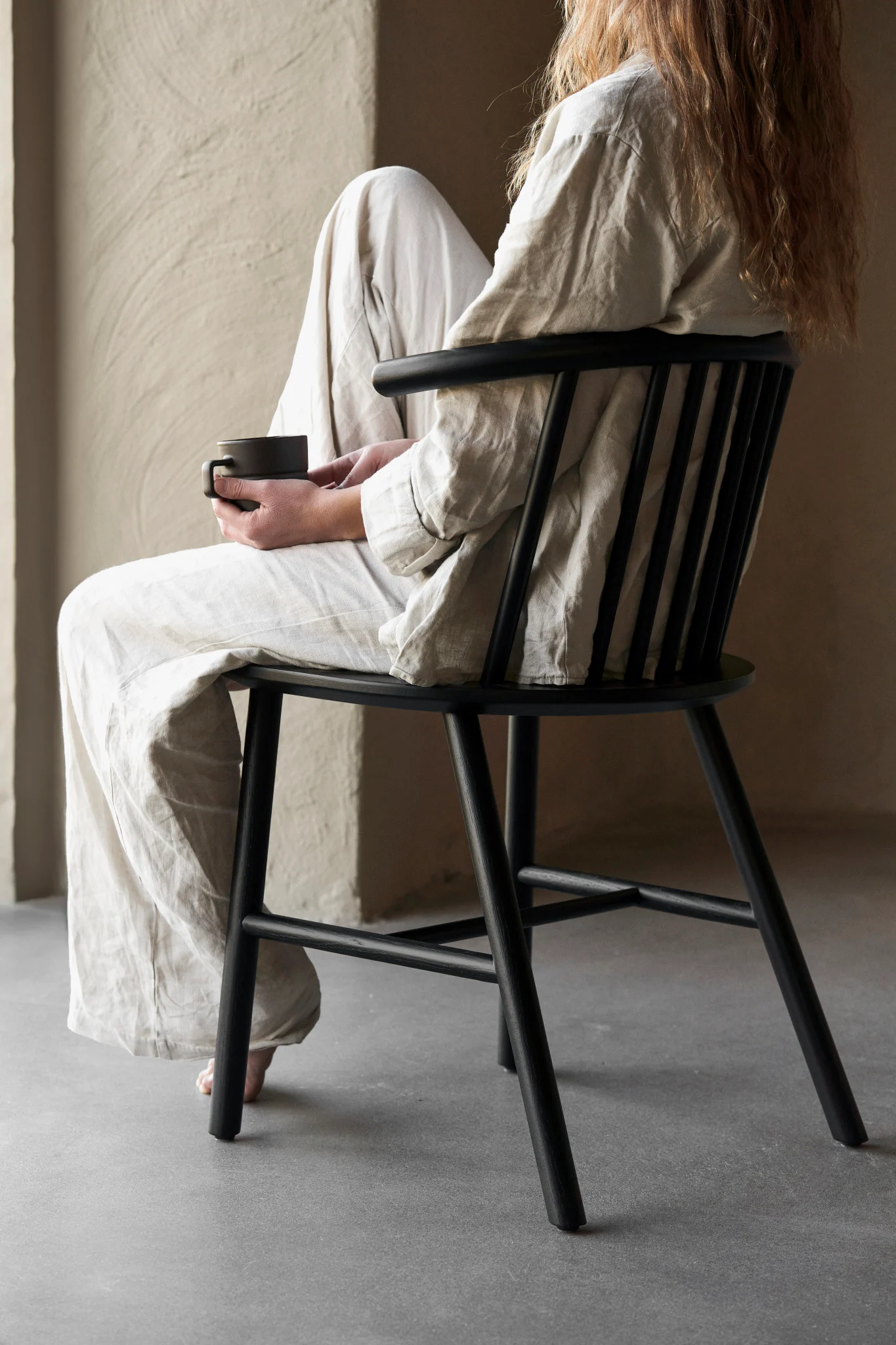 Person with long hair sitting on a black wooden chair, wearing light linen clothes and holding a dark mug.