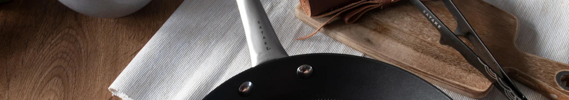 Satake pan and tongs on a wooden board and striped towel with leather accents.