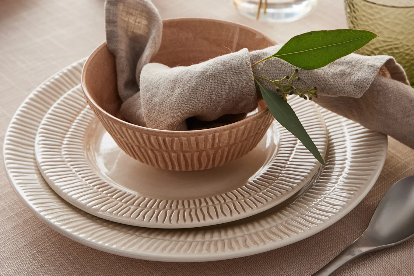Table setting with stacked textured plates, a matching bowl, a beige linen napkin, and a eucalyptus sprig.