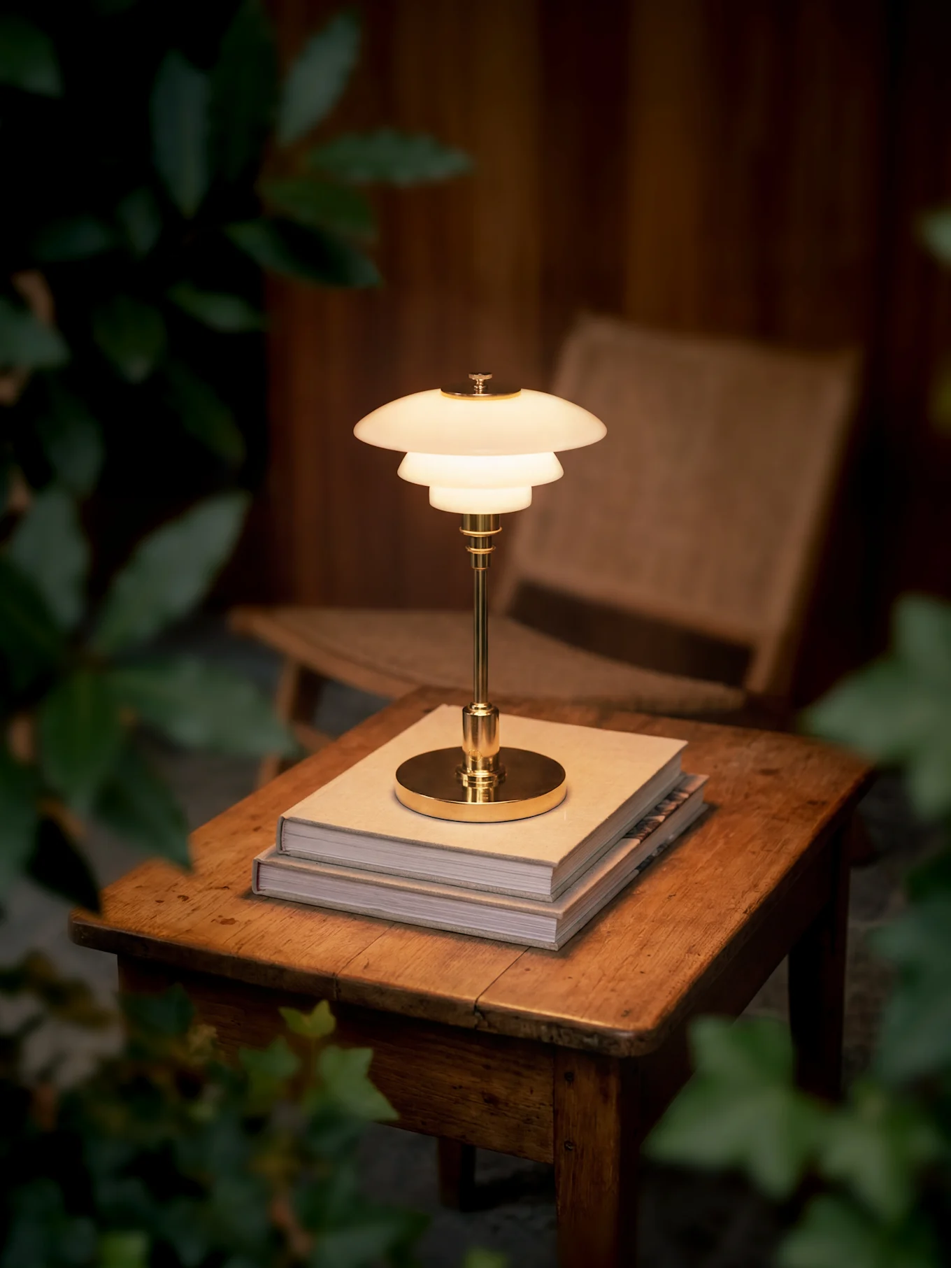 A glowing, multi-tiered brass table lamp sits on two books on a wooden table, surrounded by dark foliage.