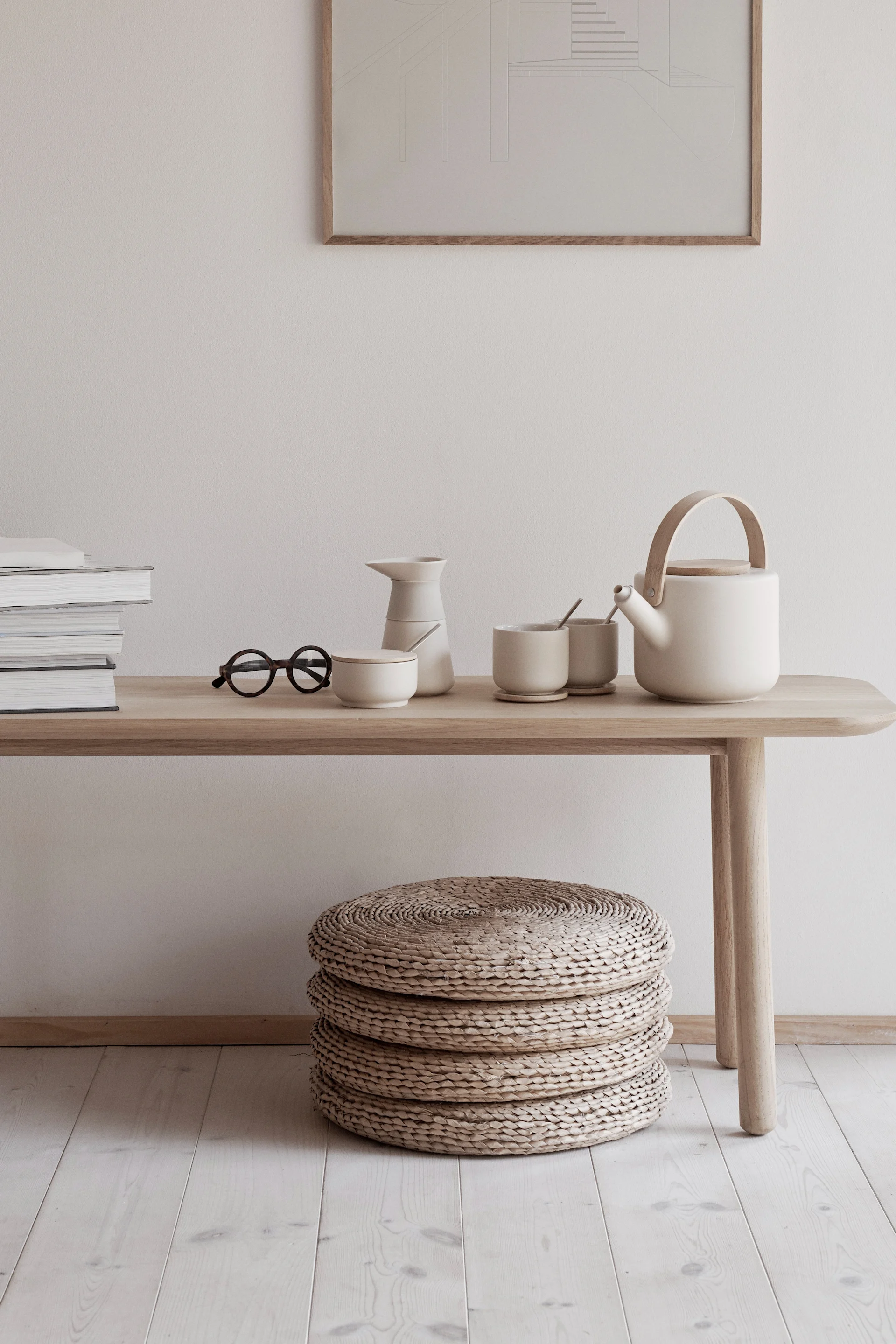 A minimalist room with a light wooden bench holding books, glasses, and a cream ceramic tea set. Three woven poufs are on the floor.