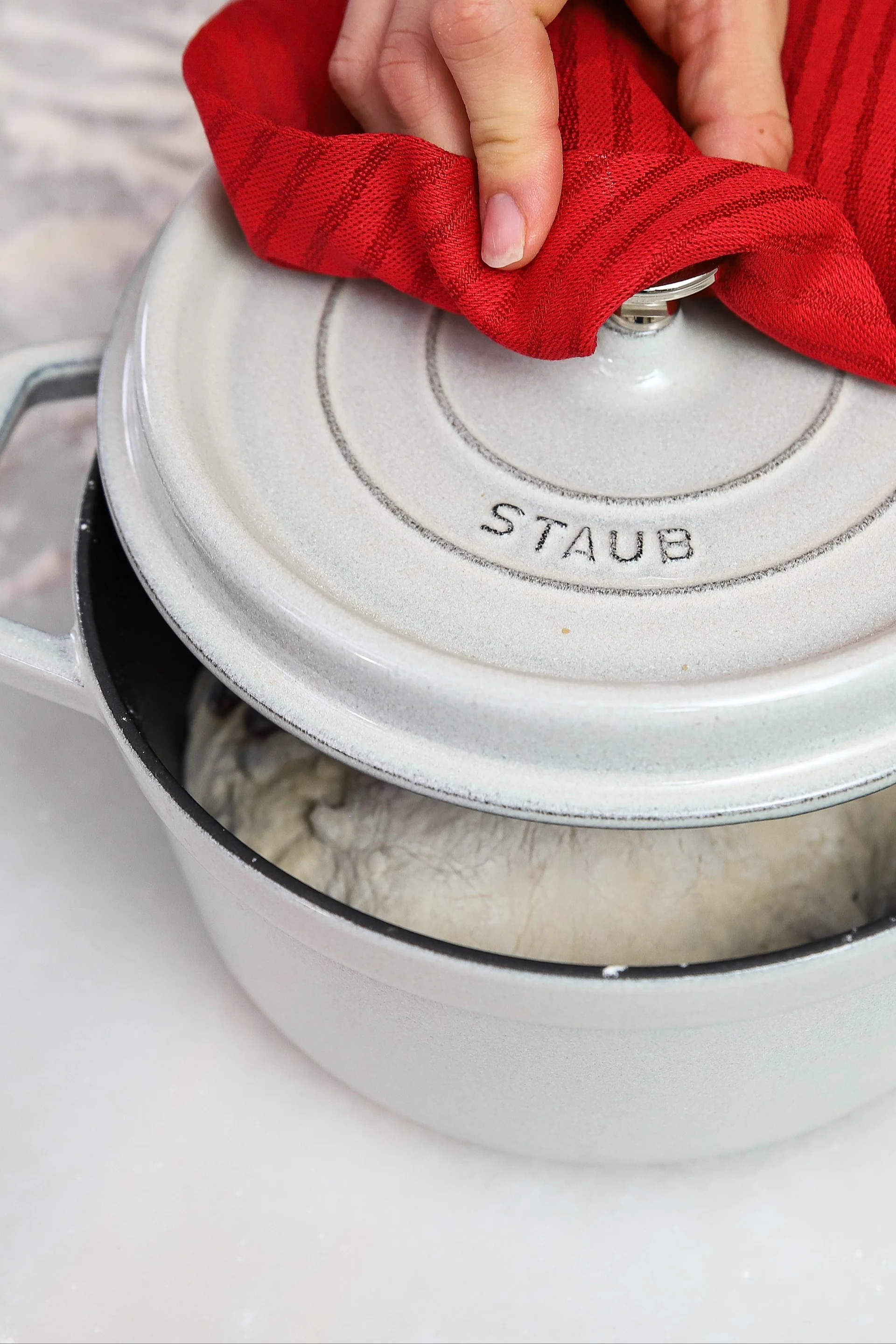 Hand in red towel lifting the lid of a white Dutch oven to reveal dough inside.