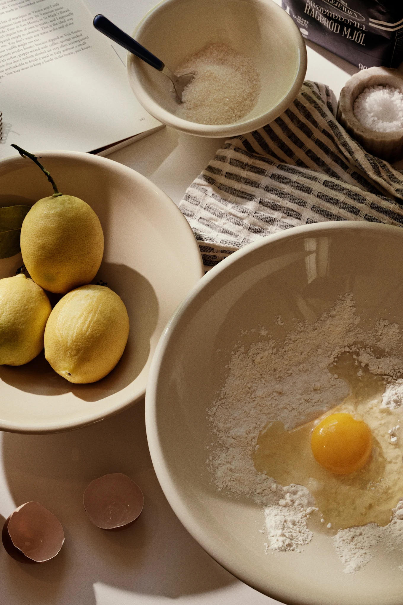 Baking ingredients: lemons in a bowl, sugar, flour with an egg yolk, and eggshells on a table.