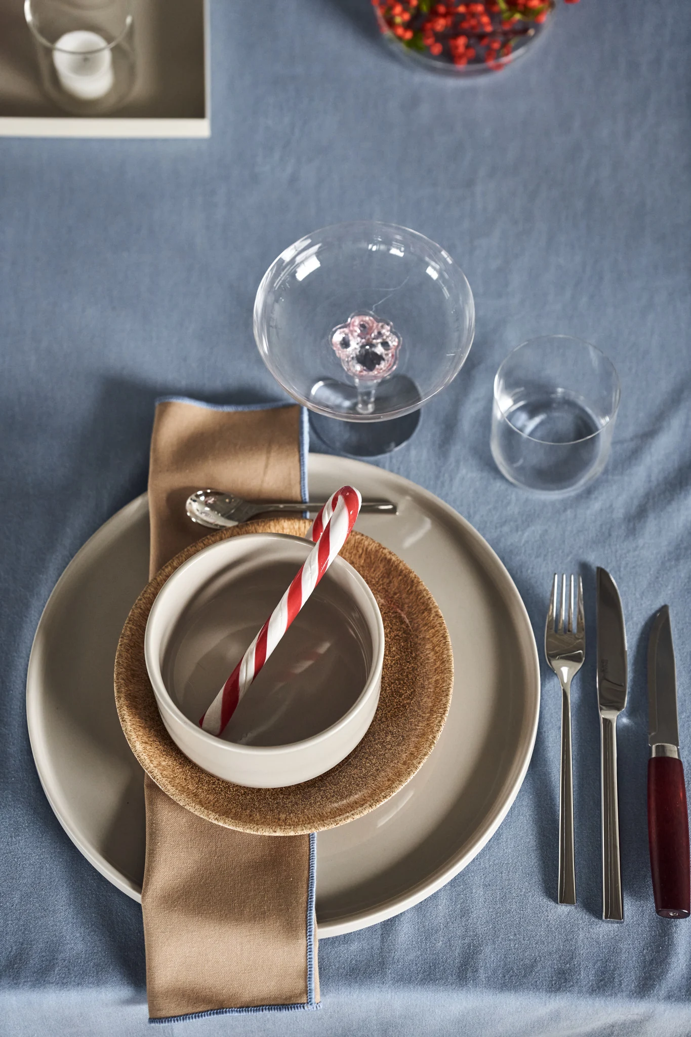 A festive table setting on a blue tablecloth with plates, a bowl holding a candy cane, cutlery, and glasses.