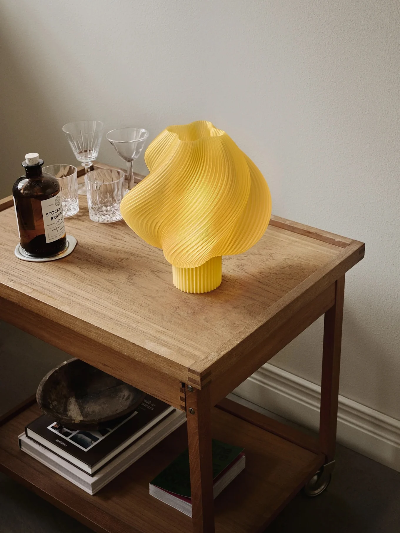 Yellow pleated lamp on a wooden rolling cart with a gin bottle, glasses, and books on the lower shelf.
