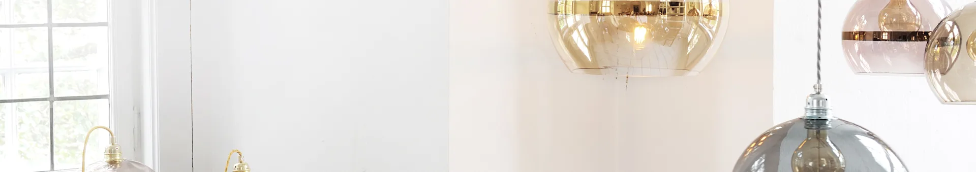 A room with a window and three hanging pendant lights: one gold-tinted, one pink with a gold band, and one smoky gray.