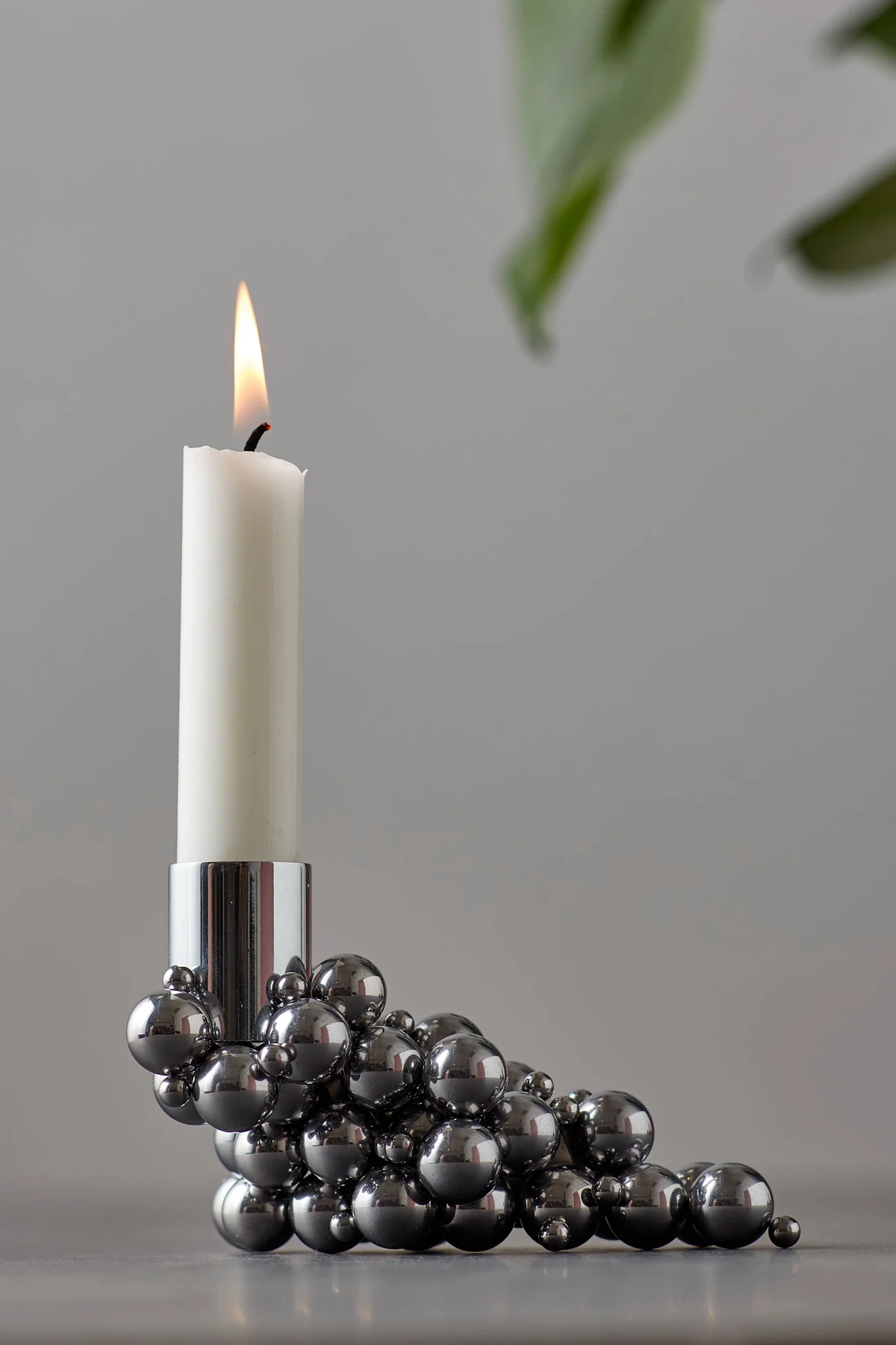 A lit white candle sits in a metallic candle holder made of many shiny silver spheres, with a blurred green leaf in the background.