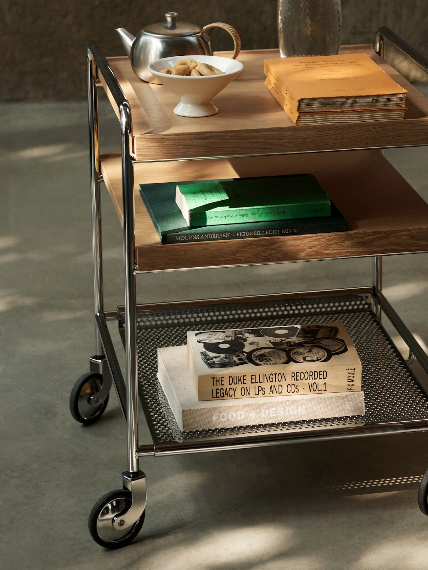 A three-tiered serving trolley with chrome frame, wood and perforated metal shelves, holding books, teapot, and cookies.