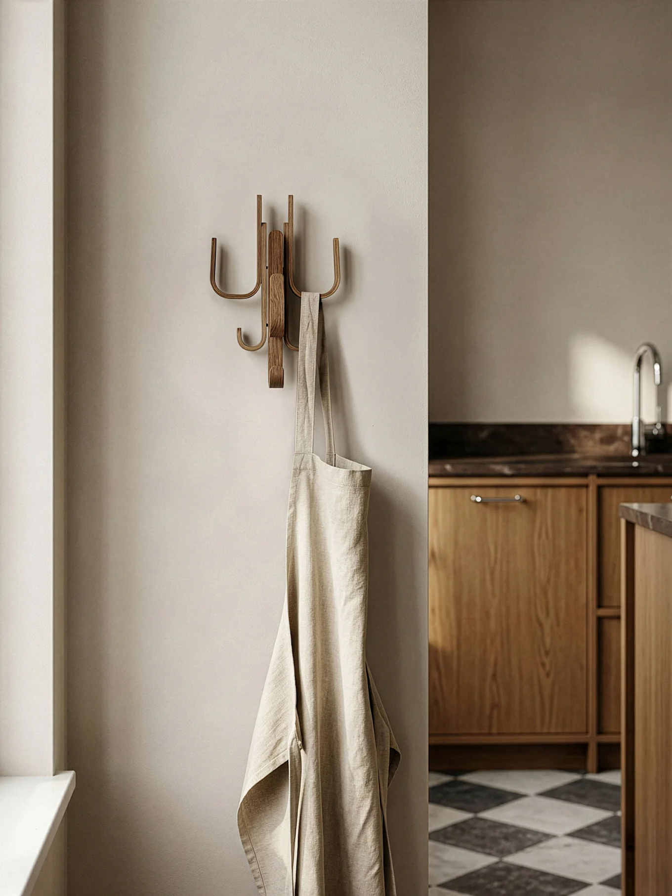A light beige linen apron hangs on a wooden wall hook in a modern kitchen with wooden cabinets and a checkered floor.