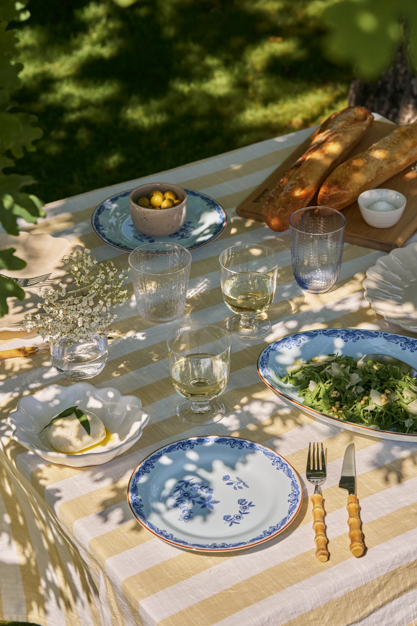 Outdoor dining table set with white and yellow striped tablecloth, blue floral plates, wine, salad, mozzarella, olives, and baguettes.
