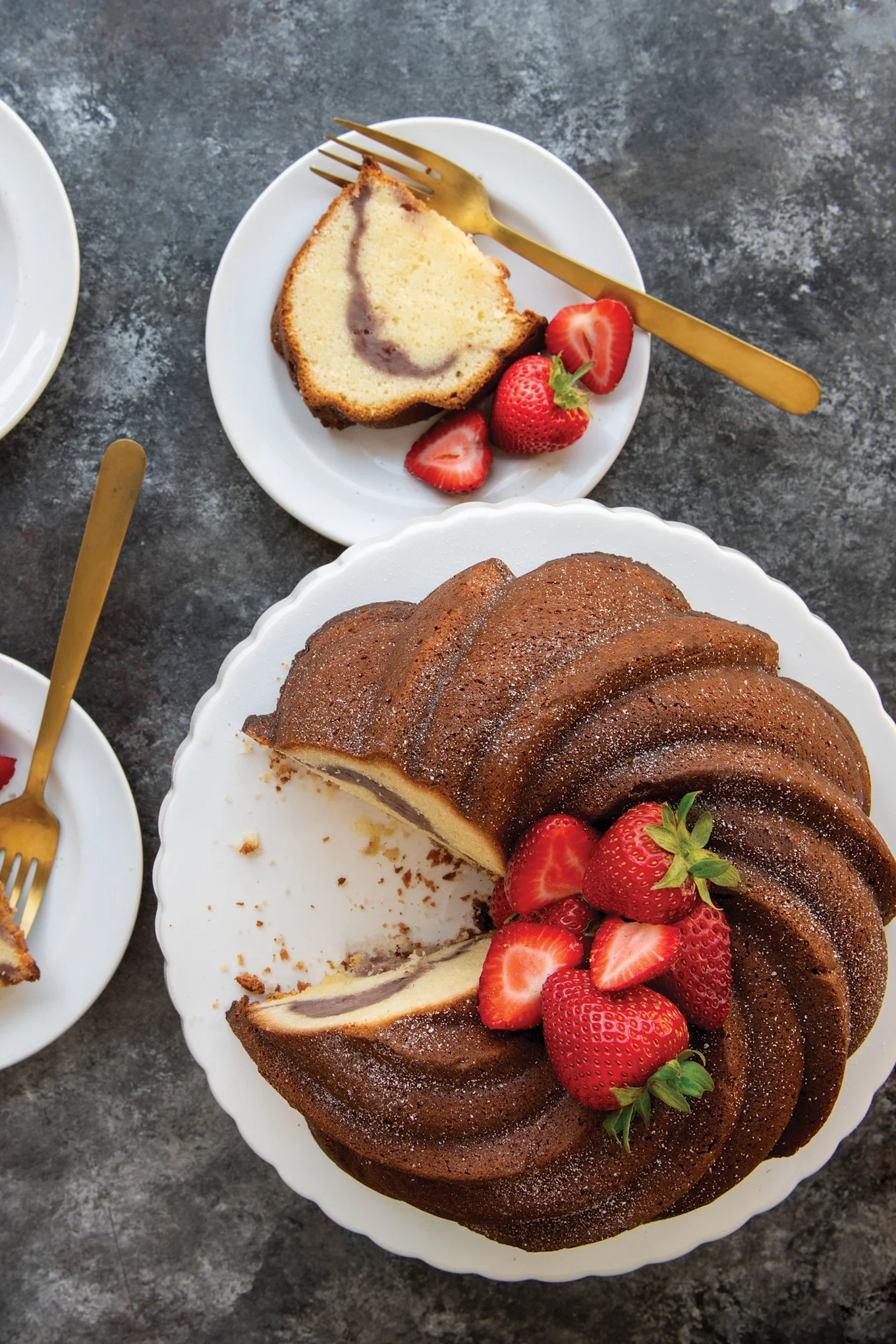 Bundt cake with a swirled interior, dusted with powdered sugar and garnished with fresh strawberries. Two slices are on white plates.