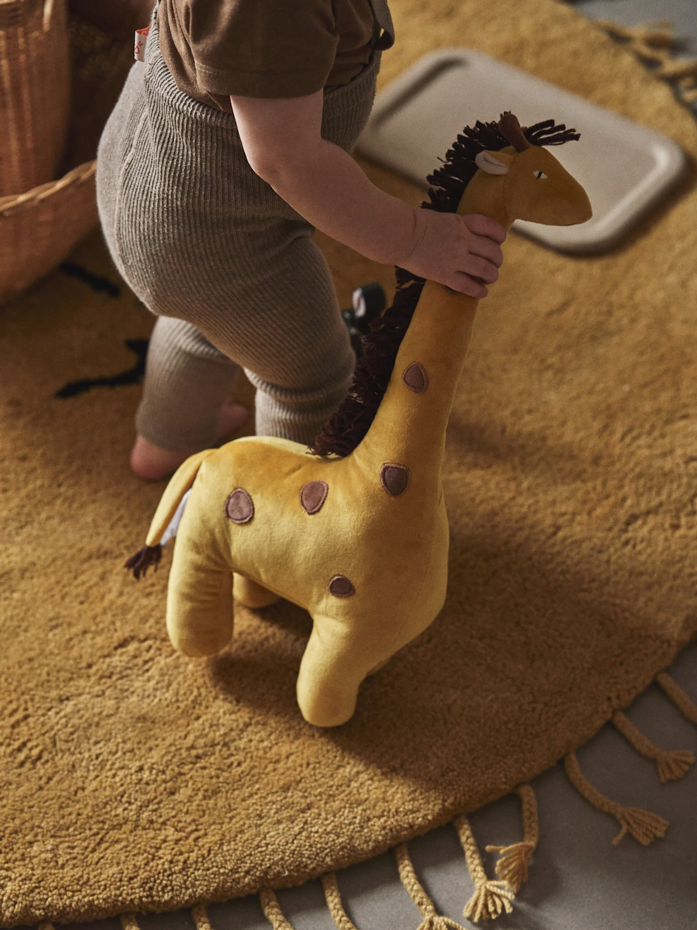 A child in ribbed overalls holds a yellow stuffed giraffe toy with brown spots on a textured yellow rug.