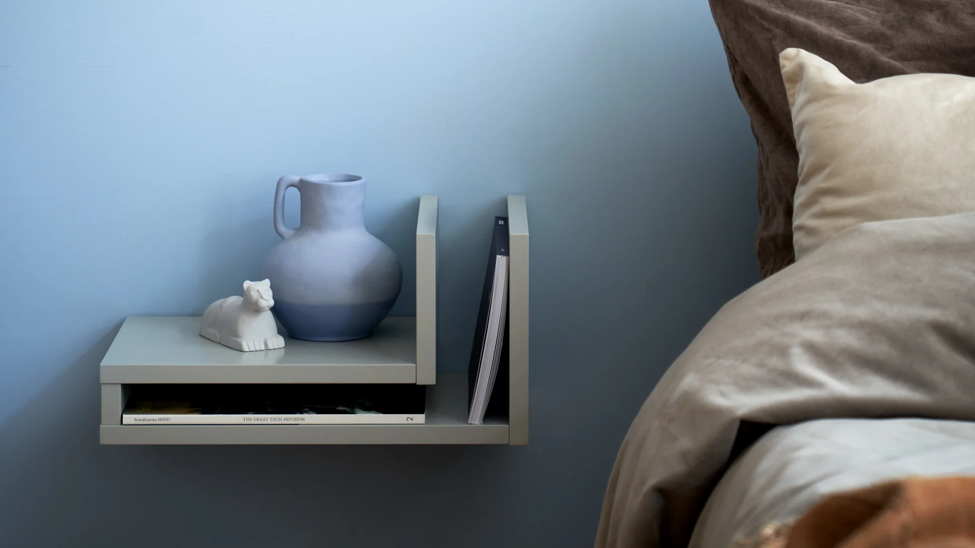 Light grey floating shelf on a blue wall with a two-toned blue vase, white animal statue, and magazines next to a bed.