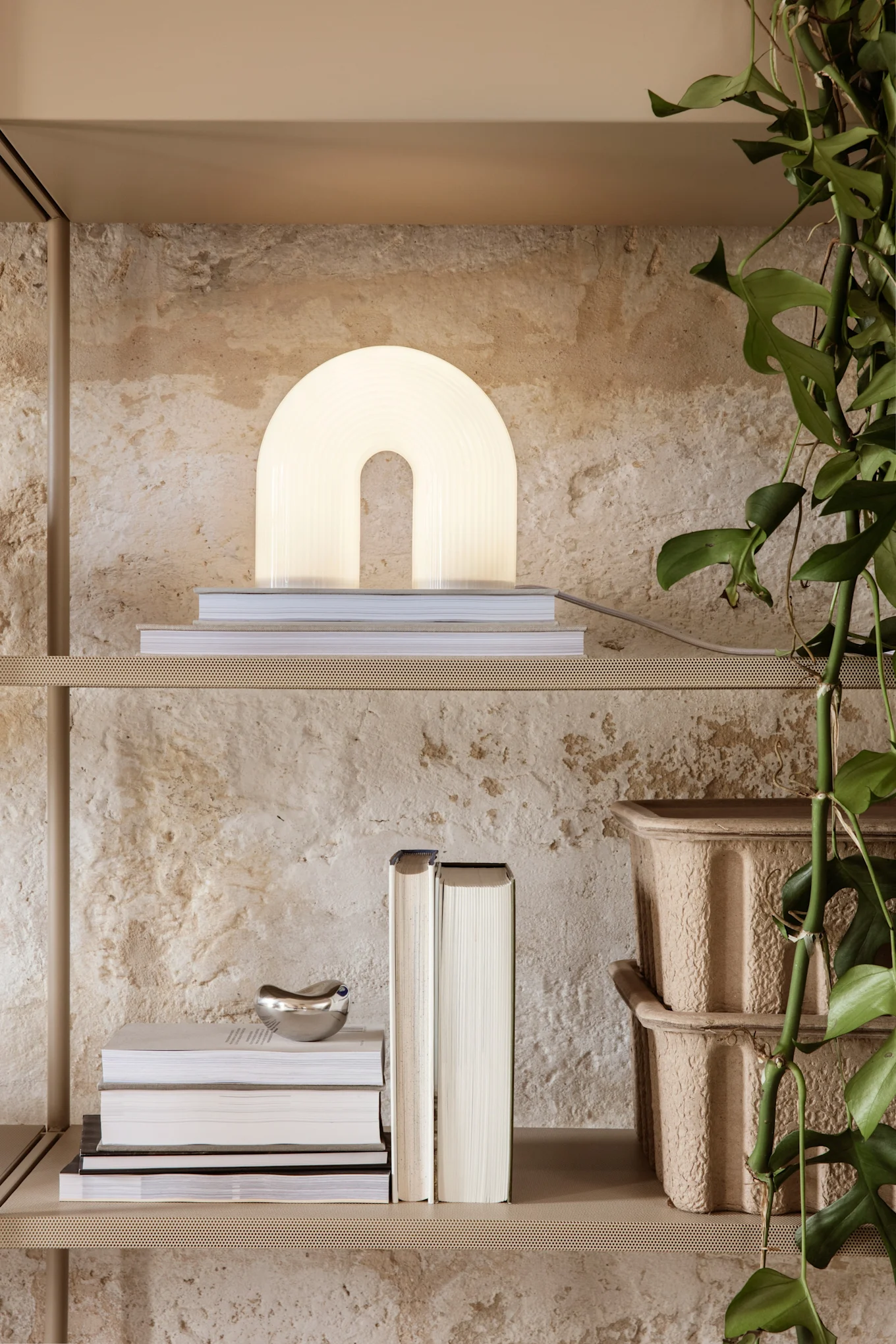 A modern arch-shaped lamp on books, with more books, silver decor, cardboard planters, and a vining plant on beige shelves.