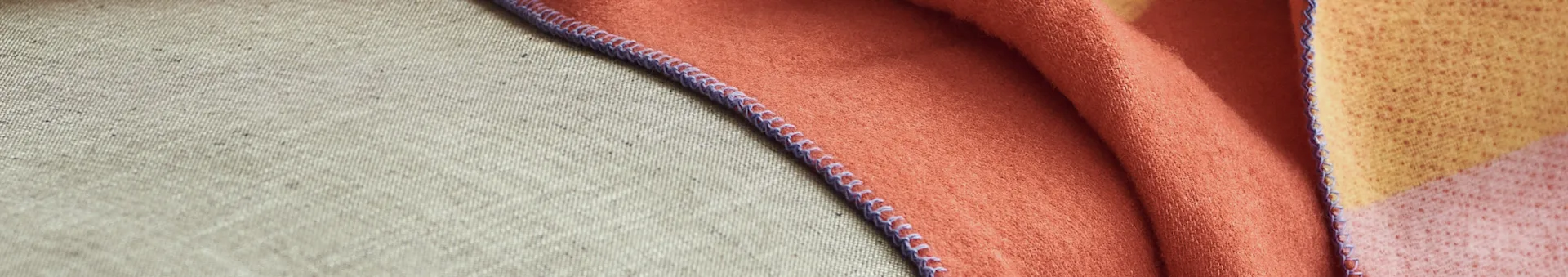 Close-up of a textured beige fabric next to a folded, soft orange, yellow, and pink blanket with a purple stitched edge.