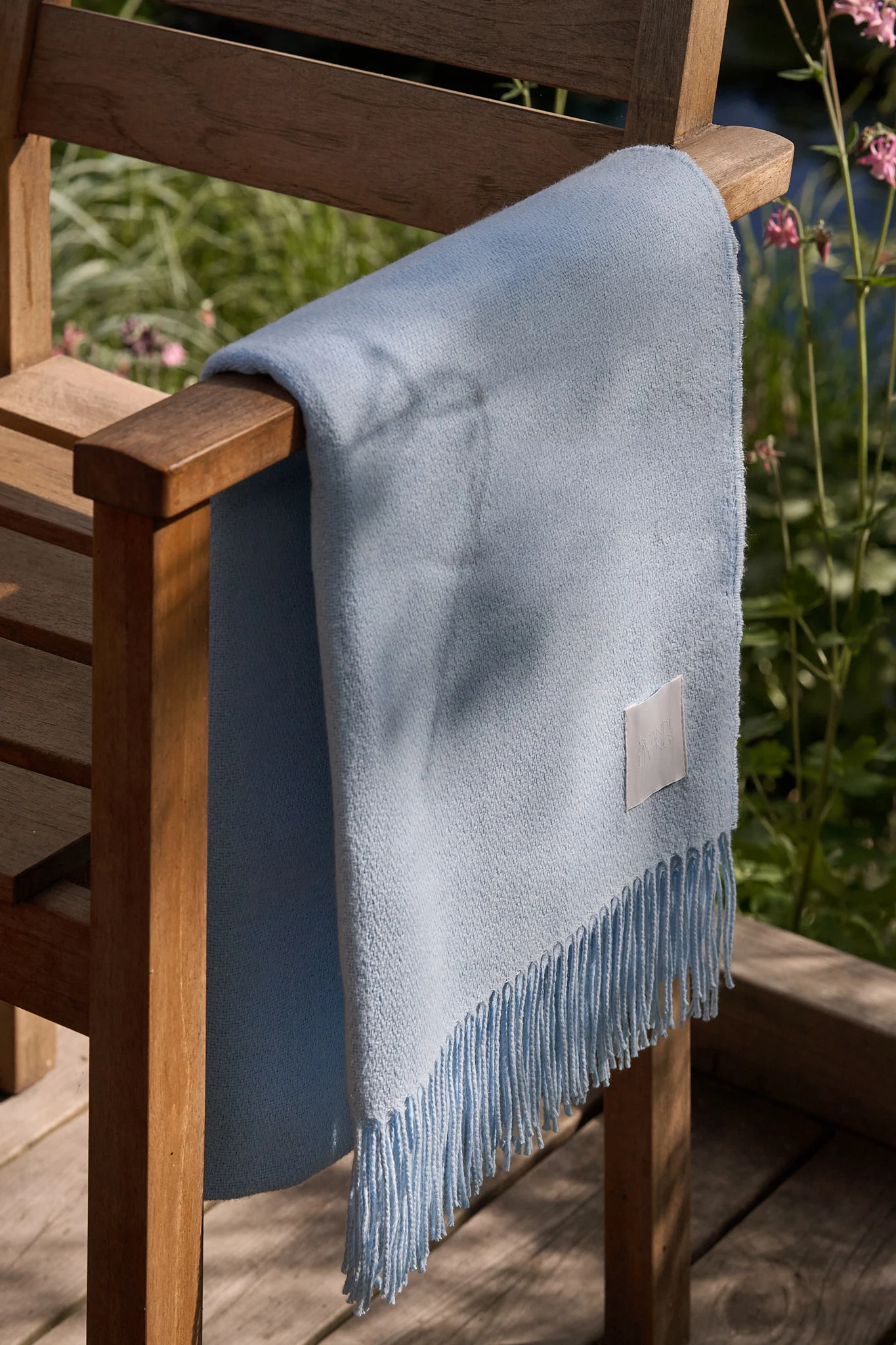 Light blue fringed blanket draped over a wooden outdoor chair in a sunny garden.