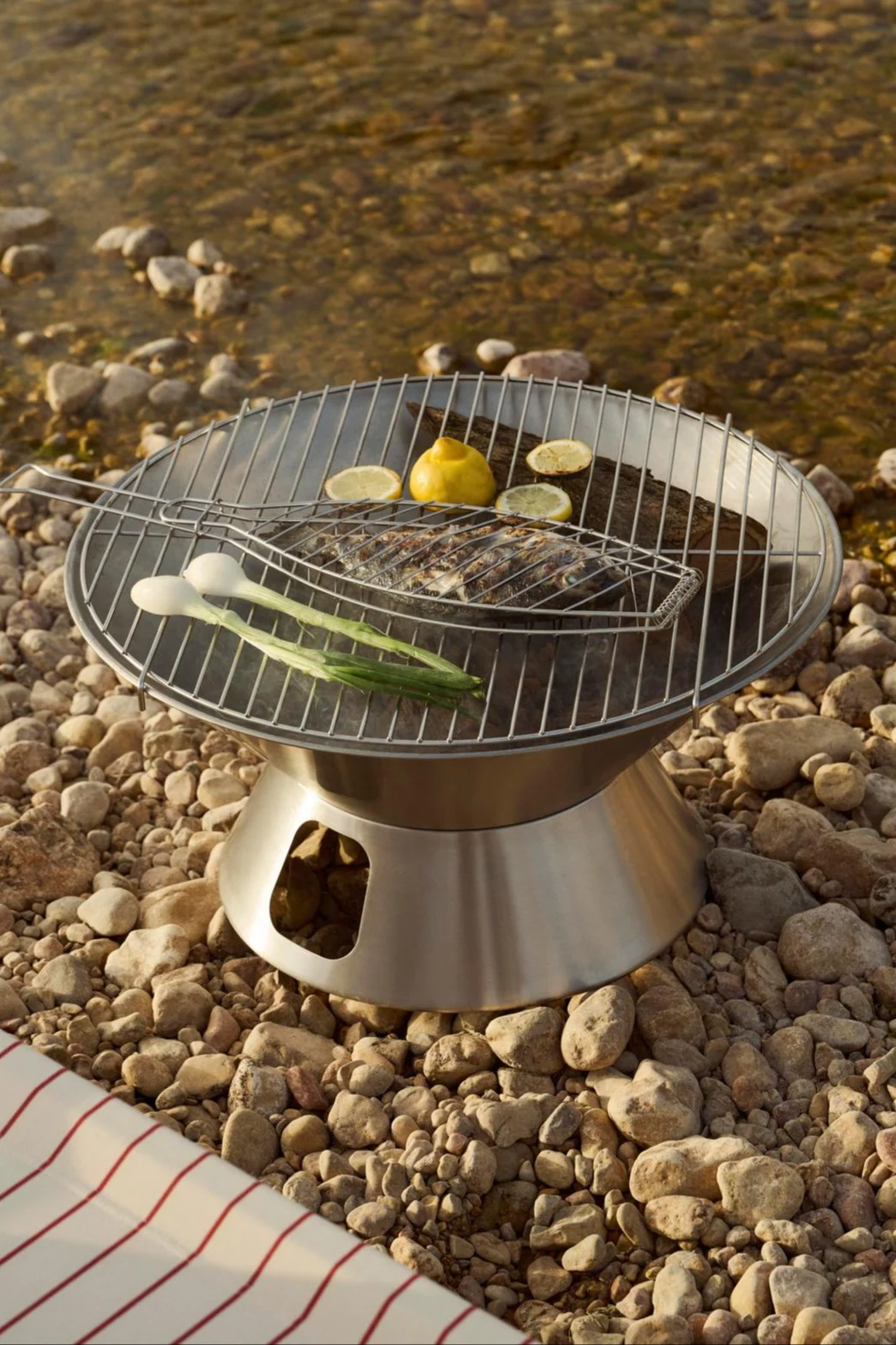 A stainless steel portable grill with a whole fish in a grill basket, lemons, and green onions on a rocky riverbank.