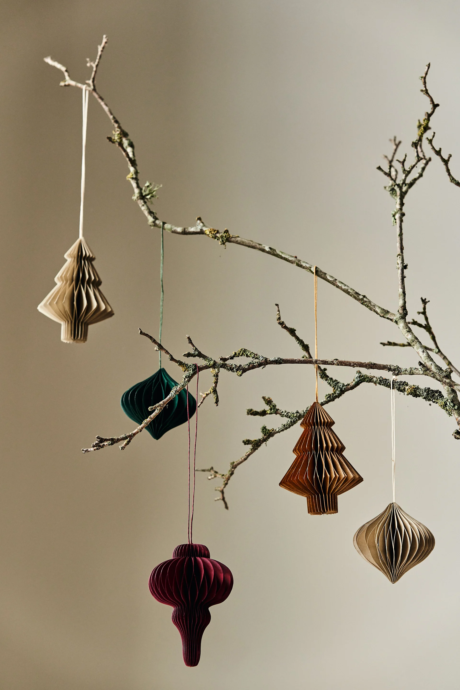 Five accordion-folded paper ornaments hanging from a mossy branch: two light brown trees, a dark green diamond, a dark red bauble, and a light brown teardrop.