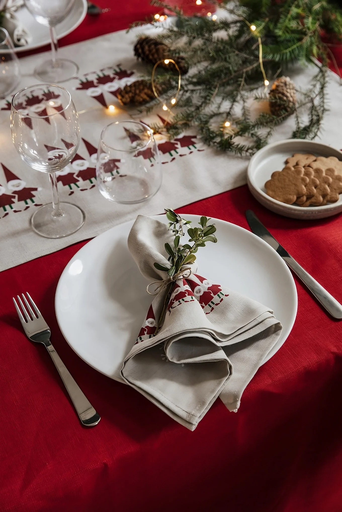 A Christmas table set with a red and gnome-patterned runner, white plate, decorated napkin, cutlery, glasses, and gingerbread.