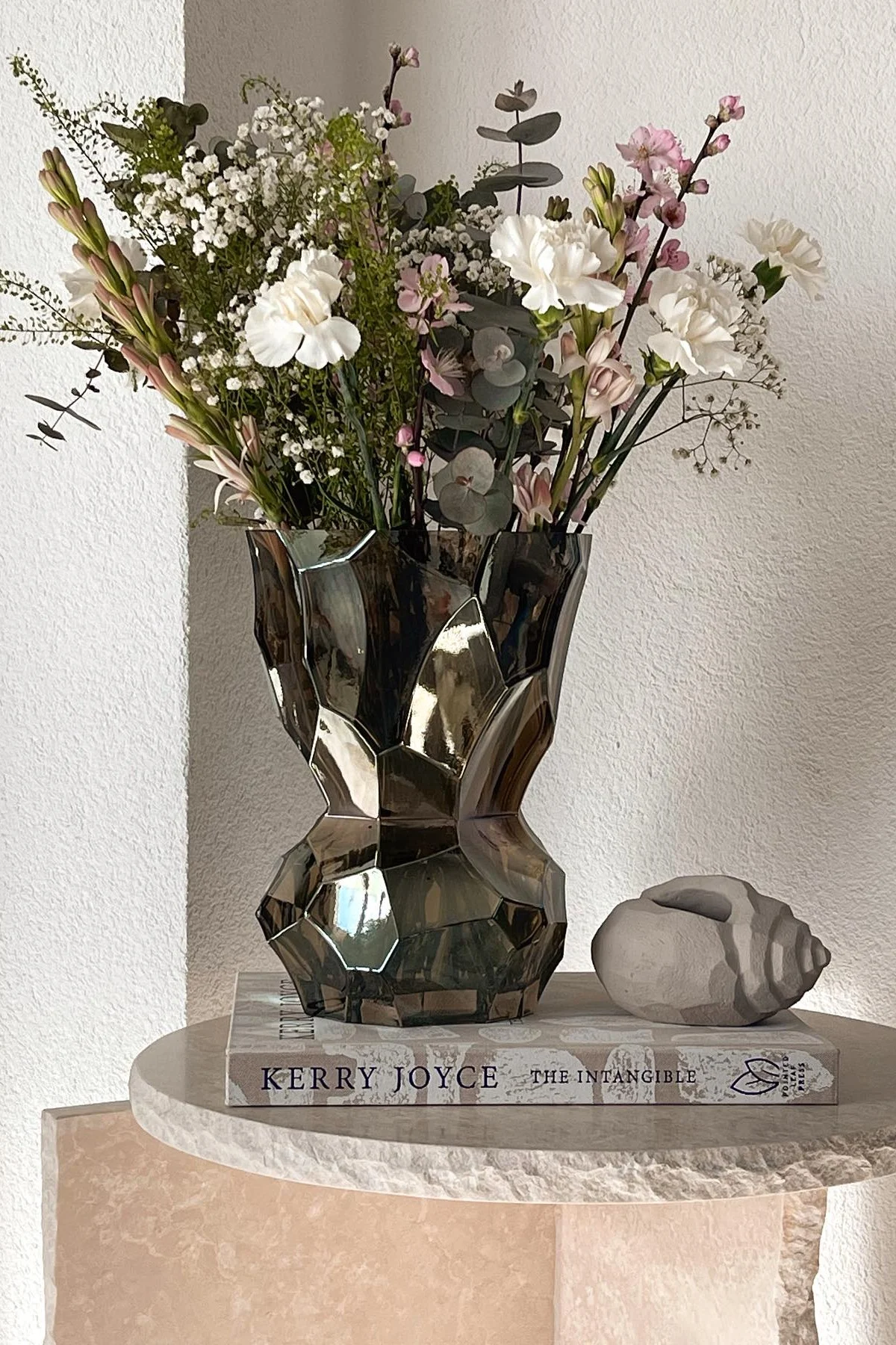A bouquet of white and pink flowers in a geometric dark vase, resting on a book and round table with a decorative shell.