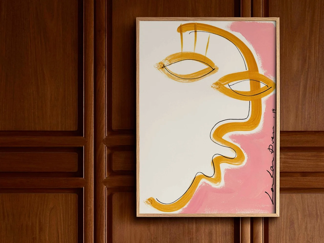 Framed abstract art print featuring a golden yellow stylized face outline on a white background with a pink section, on a paneled wooden wall.