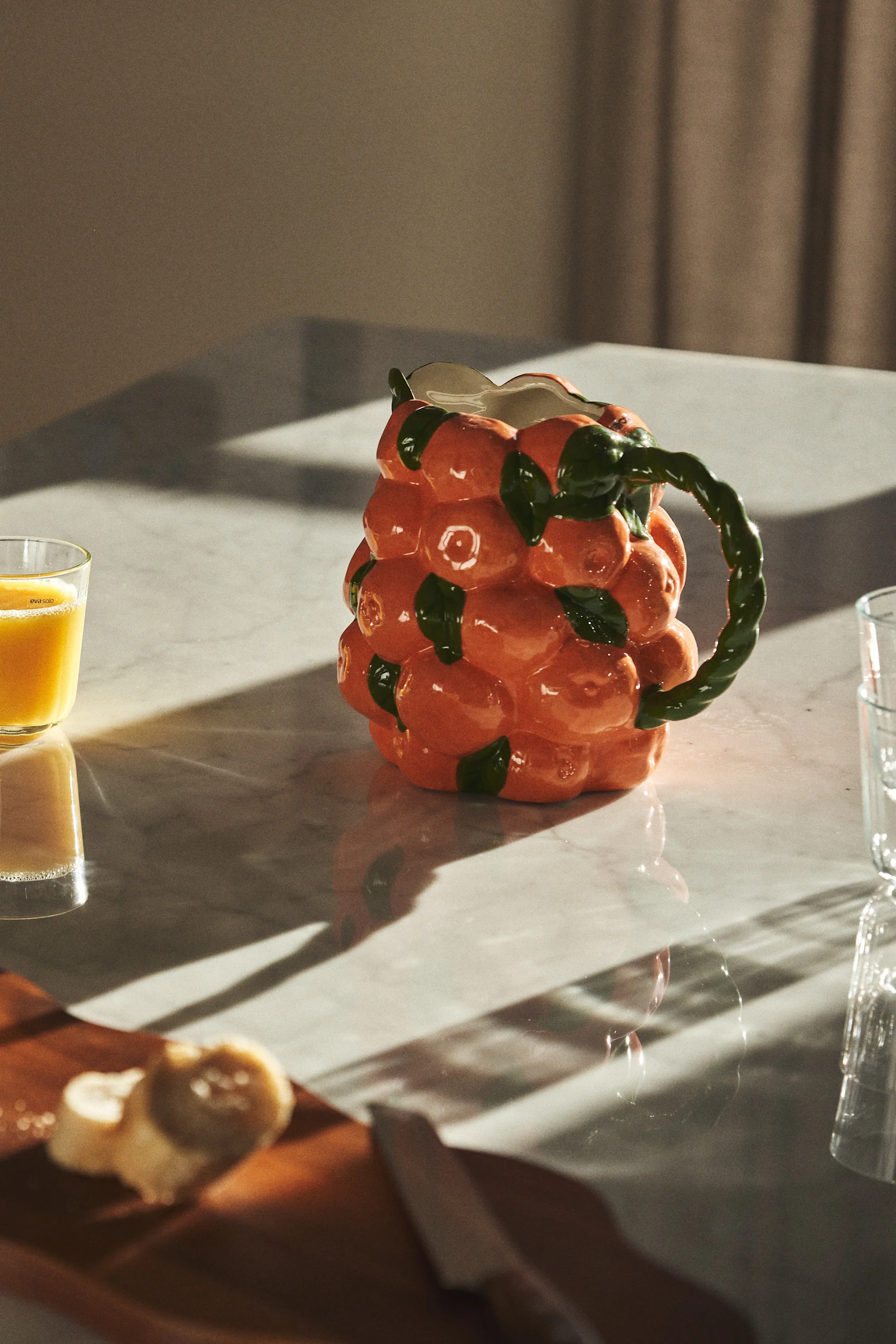 Orange ceramic jug shaped like stacked oranges with a green leafy handle, on a marble table.