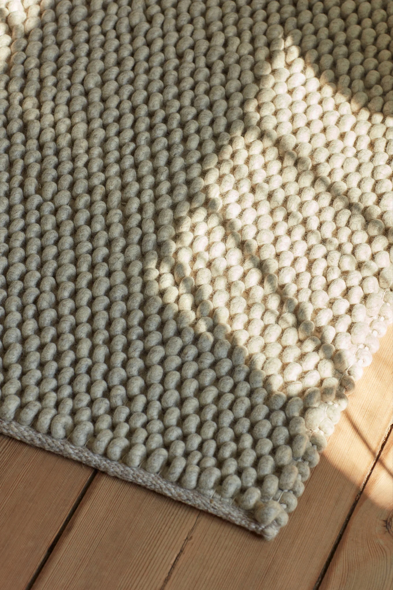 Textured rug with woven light gray wool balls on a wooden floor, with sunlight casting a diagonal shadow.