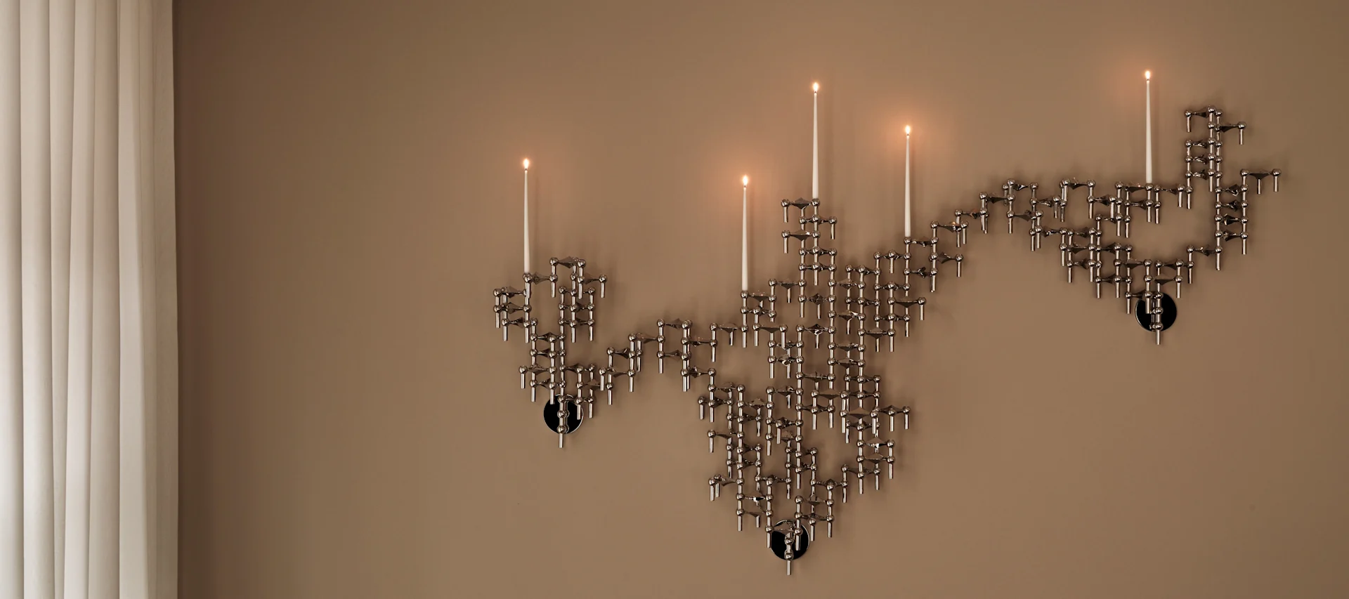 A wall-mounted modular silver-toned candelabra with several lit candles against a beige wall, beside vertical blinds.