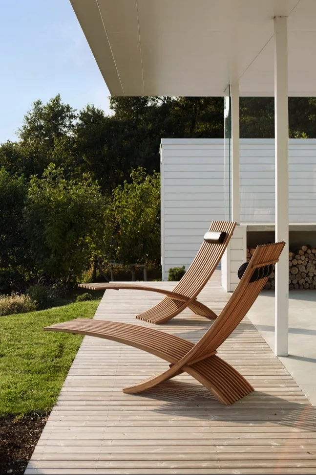 Two slatted wooden lounge chairs on a deck, with green grass and a modern white building in the background.