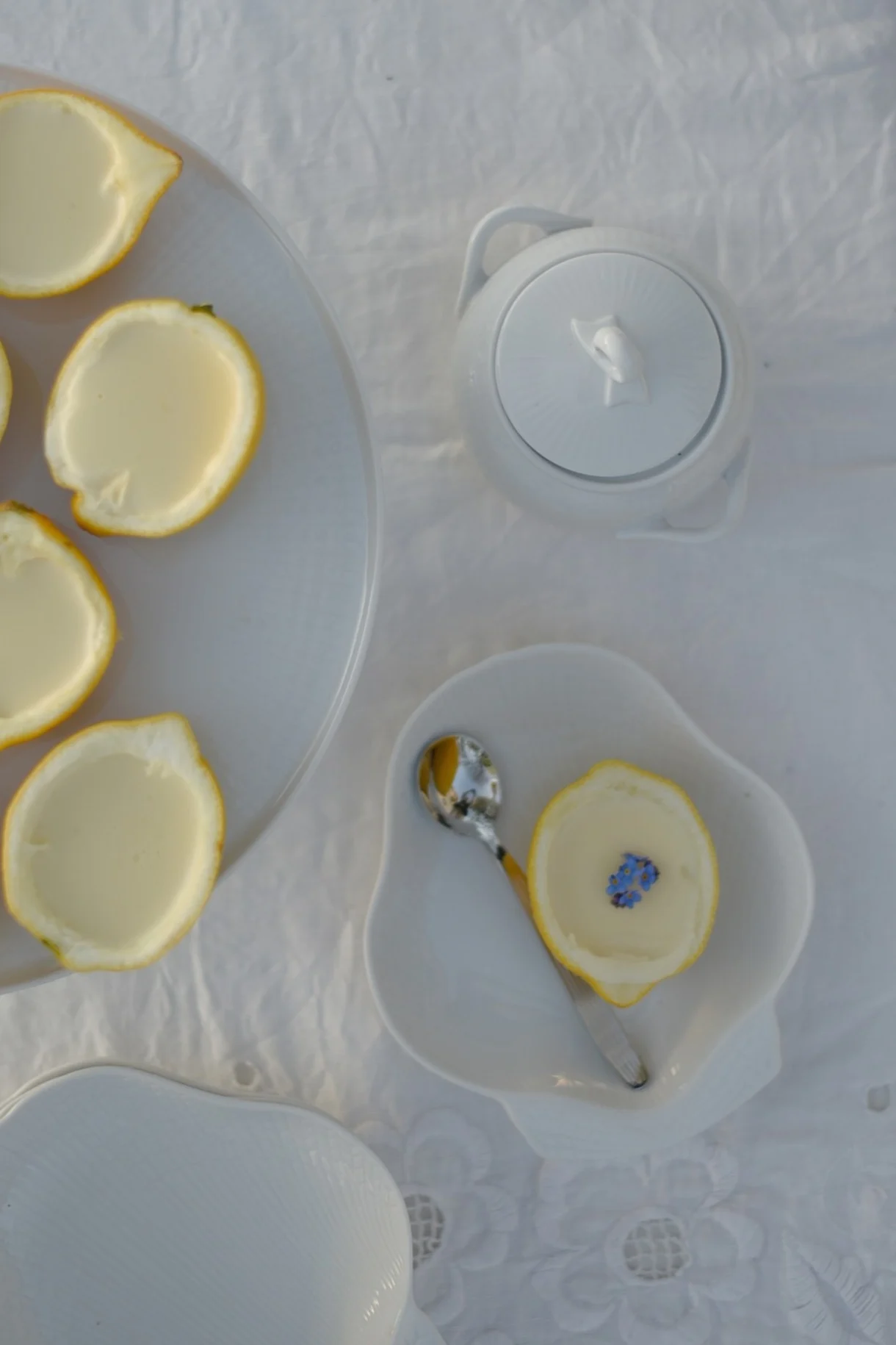Lemon halves filled with creamy dessert, one garnished with blue flowers, on white tableware with a spoon and sugar bowl.
