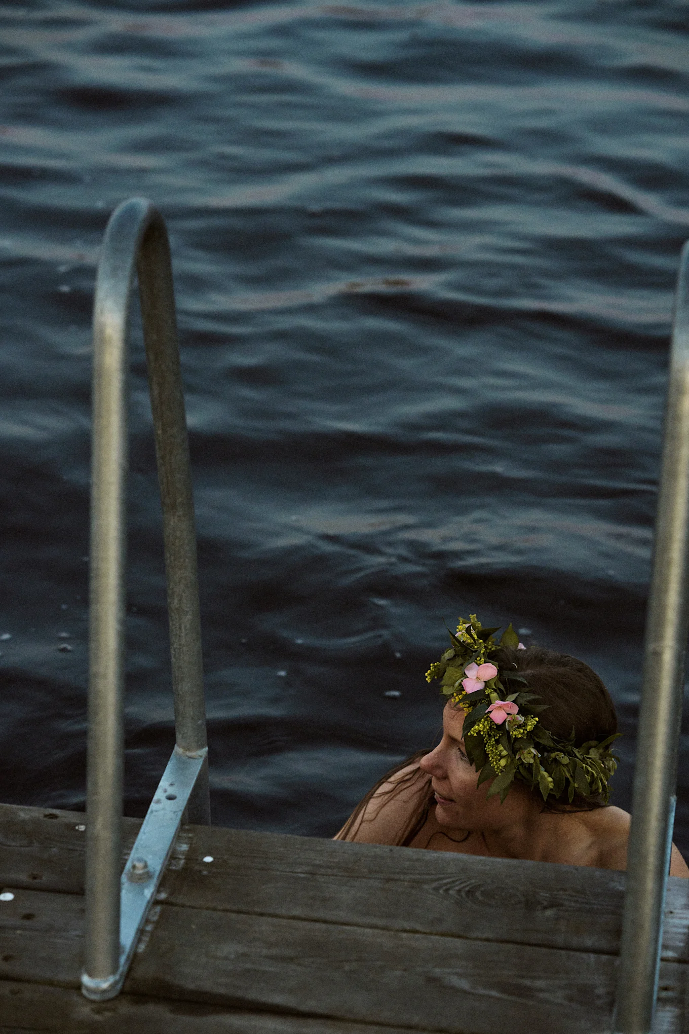 Woman in dark water wearing a flower crown, next to a wooden dock with metal handrails.