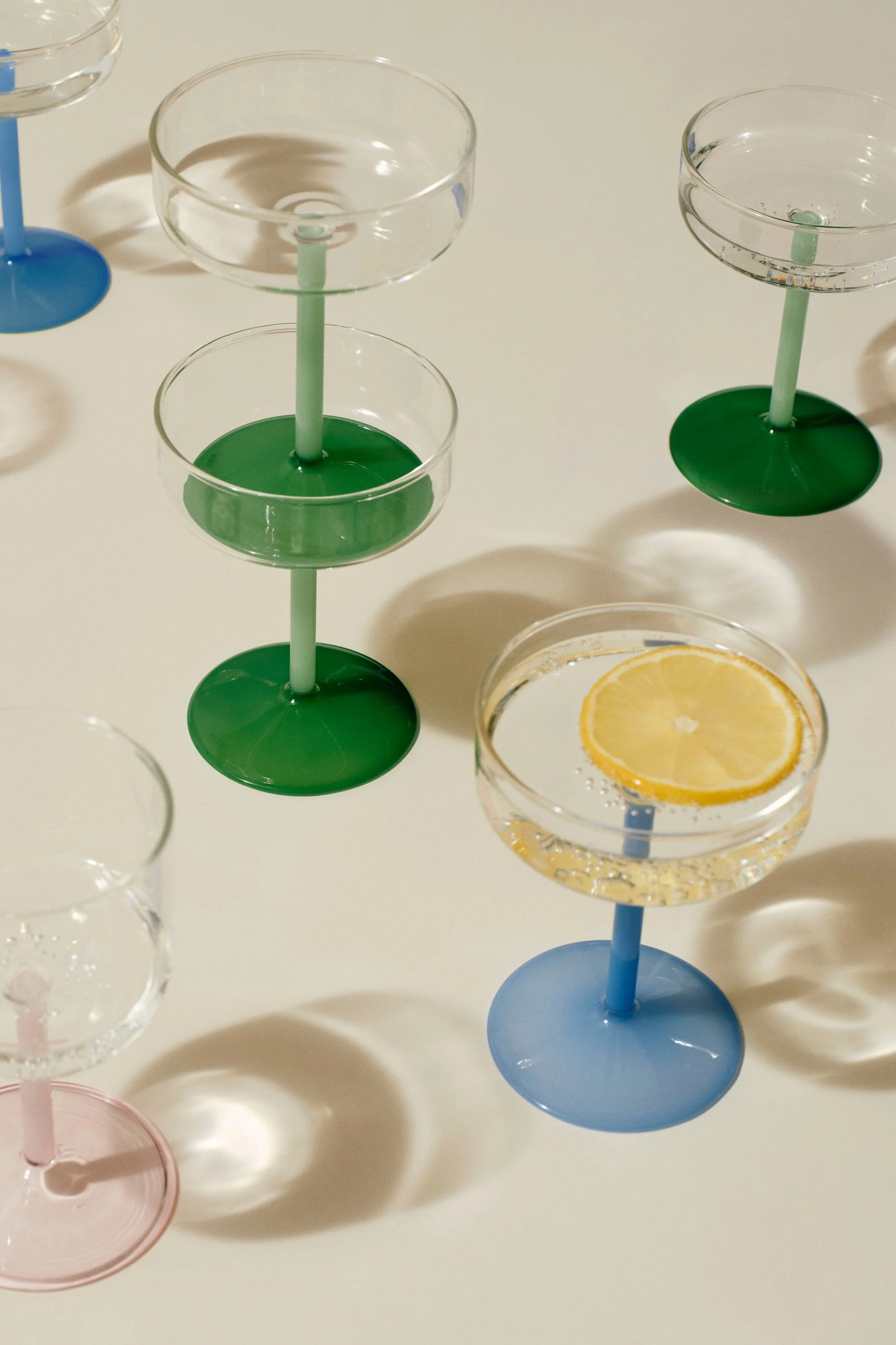 Assortment of coupe glasses with colorful stems and bases. One glass holds a lemon slice in sparkling liquid.