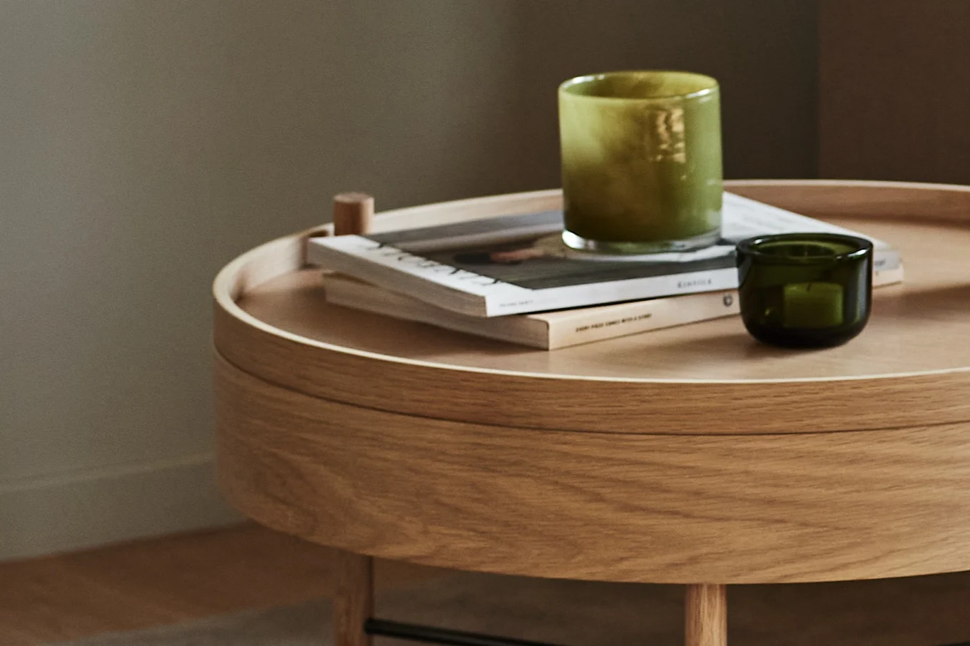 Light wood round side table with two green glass candle holders and stacked magazines.