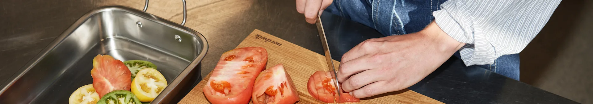 Person's hands slicing red tomatoes on a wooden cutting board, with various sliced tomatoes in a metal tray.