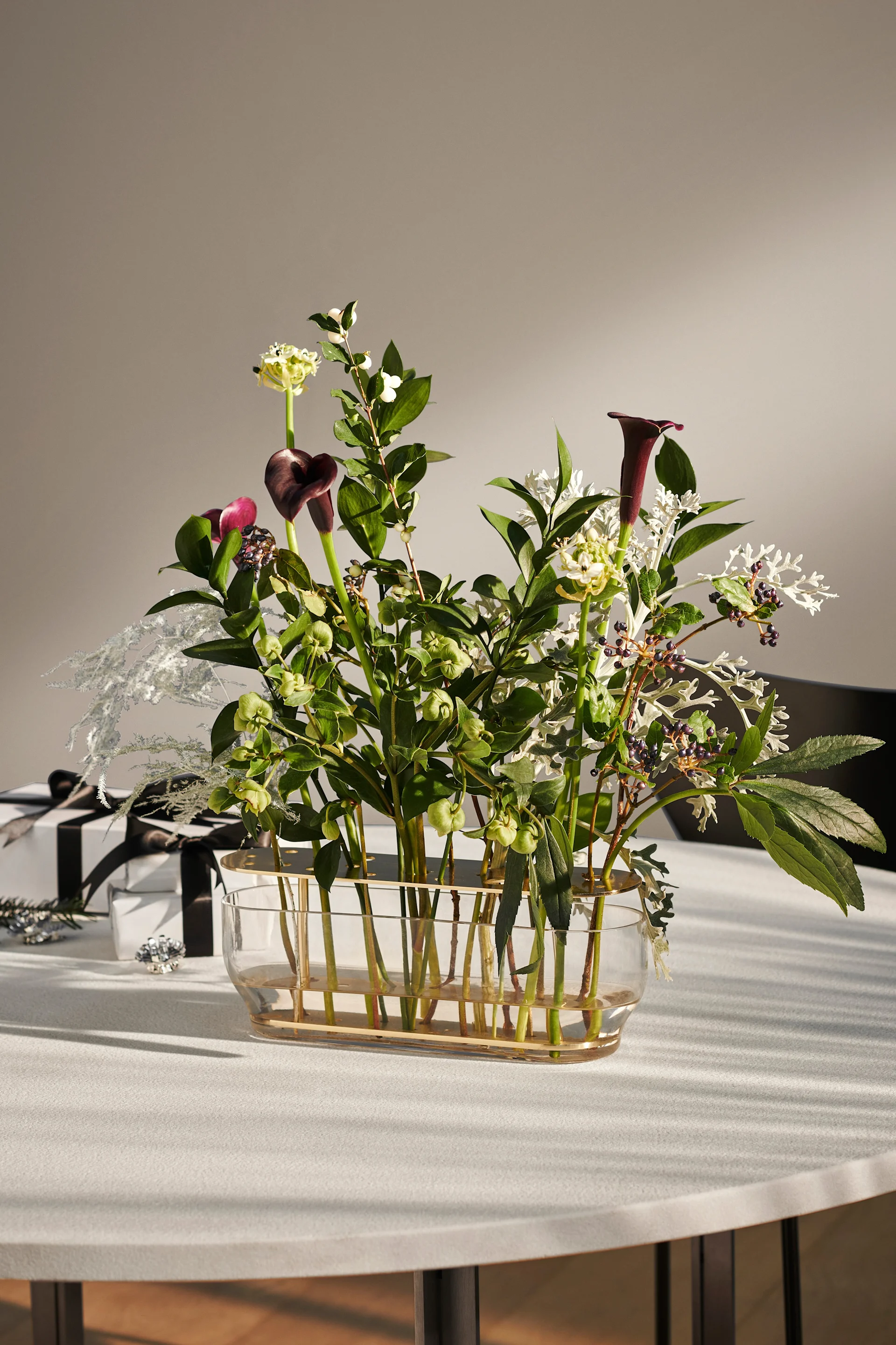 Modern floral arrangement with dark purple calla lilies, green flowers, white berries, silvery foliage in glass vase; gifts beside.