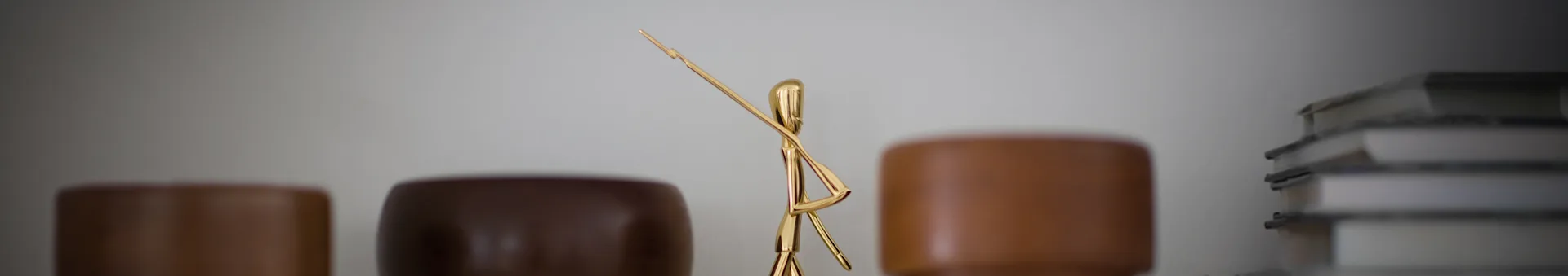 Golden abstract sculpture of a figure holding a long rod, flanked by blurred brown objects and books.