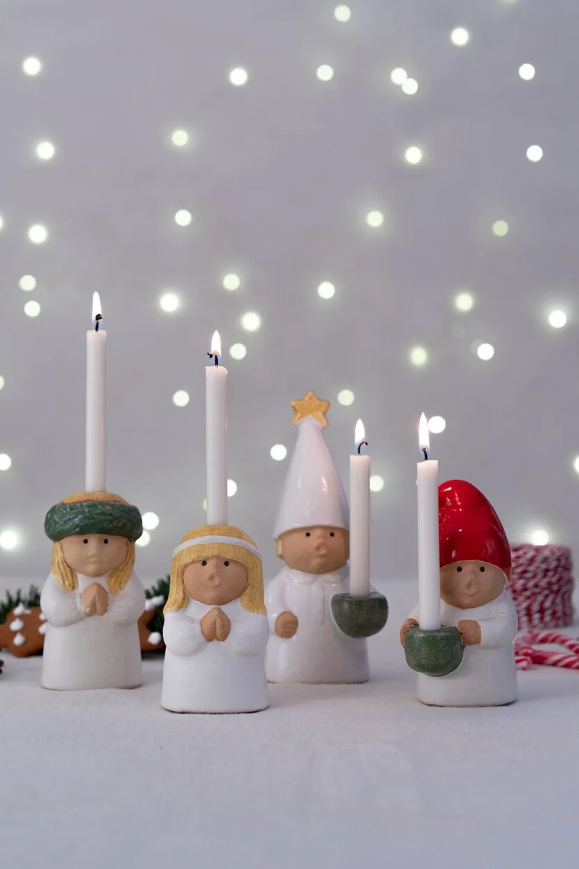 Four white ceramic figure candle holders with lit candles. Two blonde figures with clasped hands; two figures with conical hats, one red, one white with star.