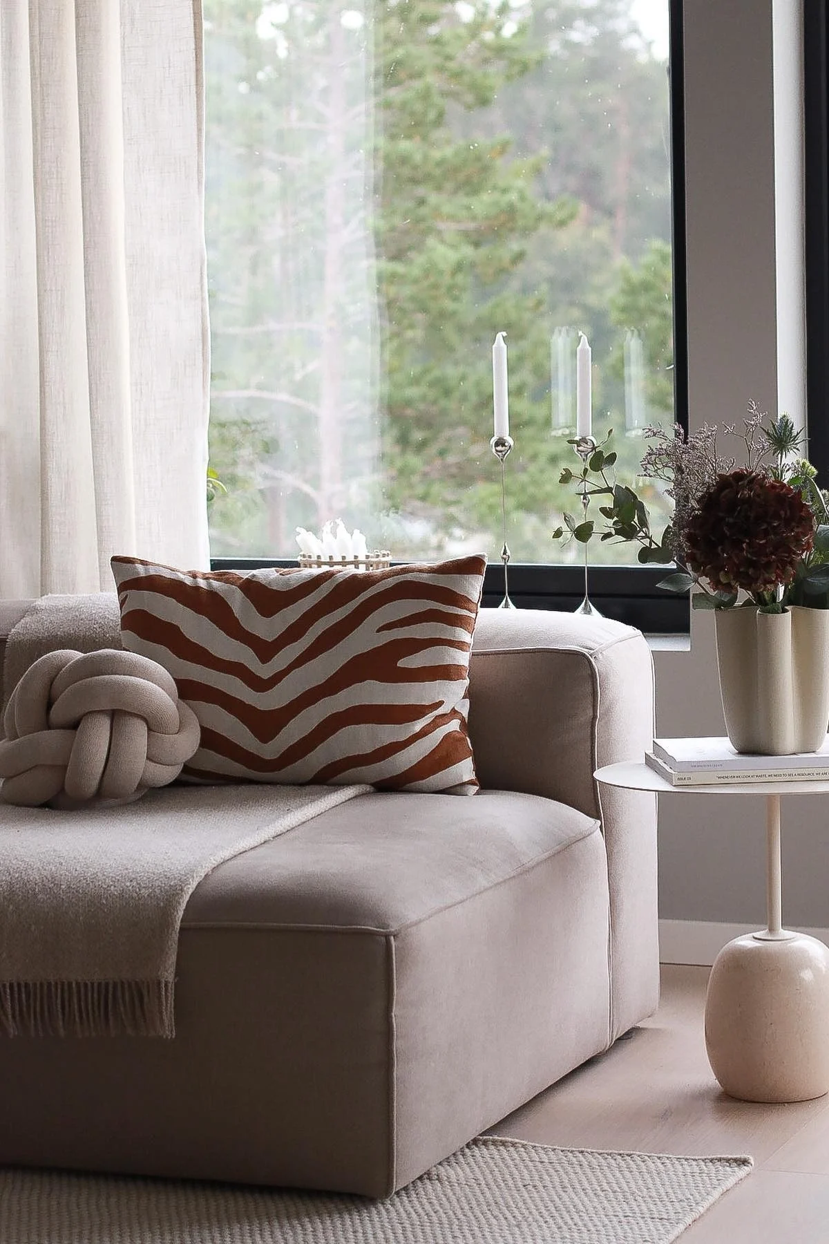 A cozy living room scene with a beige modular sofa, a knot pillow, and a zebra-print pillow by a window overlooking a forest.