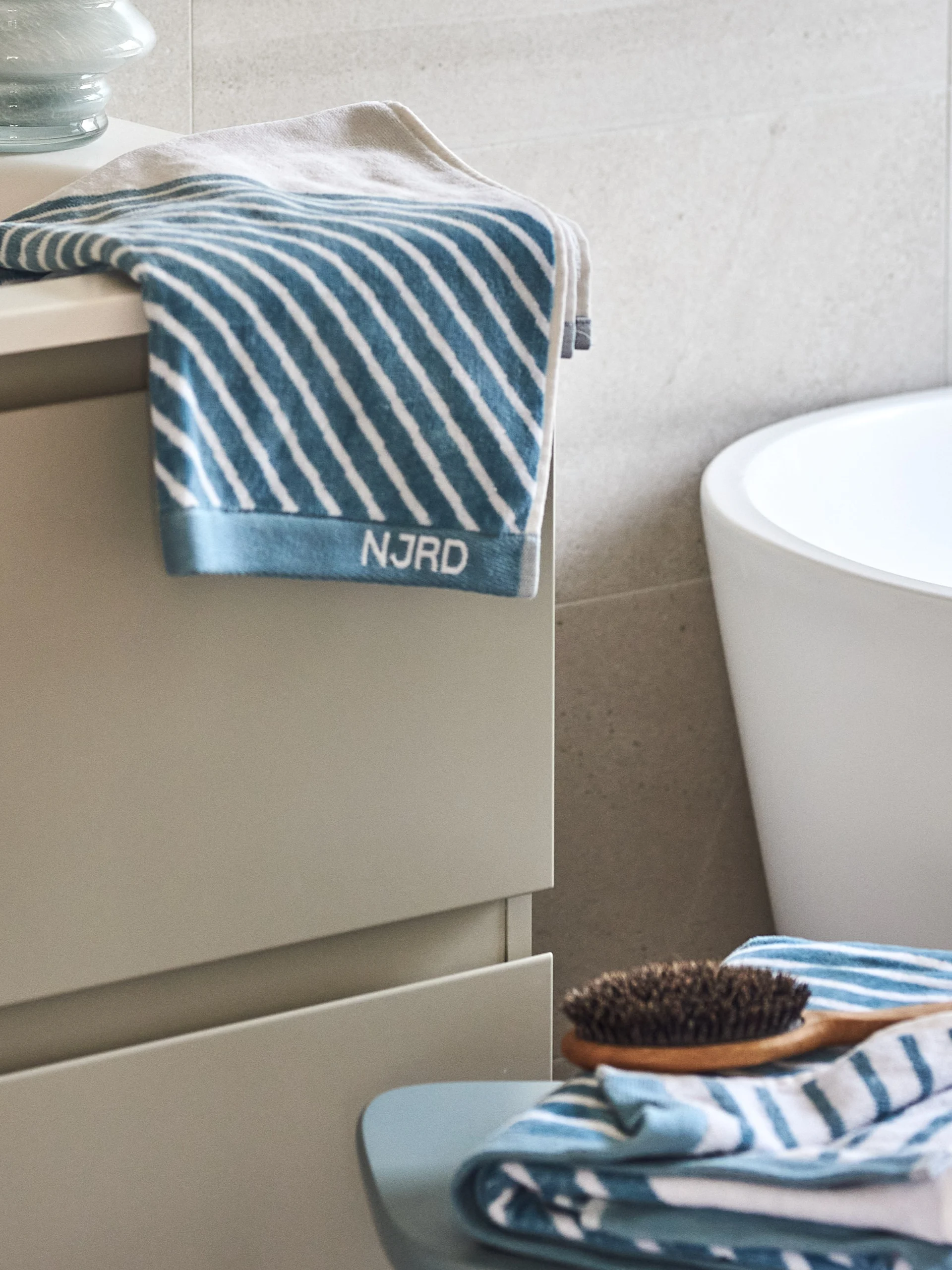Blue and white striped towels and a wooden hairbrush in a bathroom with a white tub and grey cabinet.