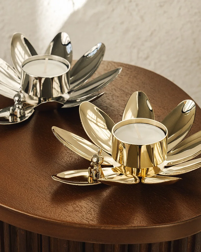 Two lotus-shaped tealight candle holders, one silver and one gold, with small Buddha figures, on a wooden table.