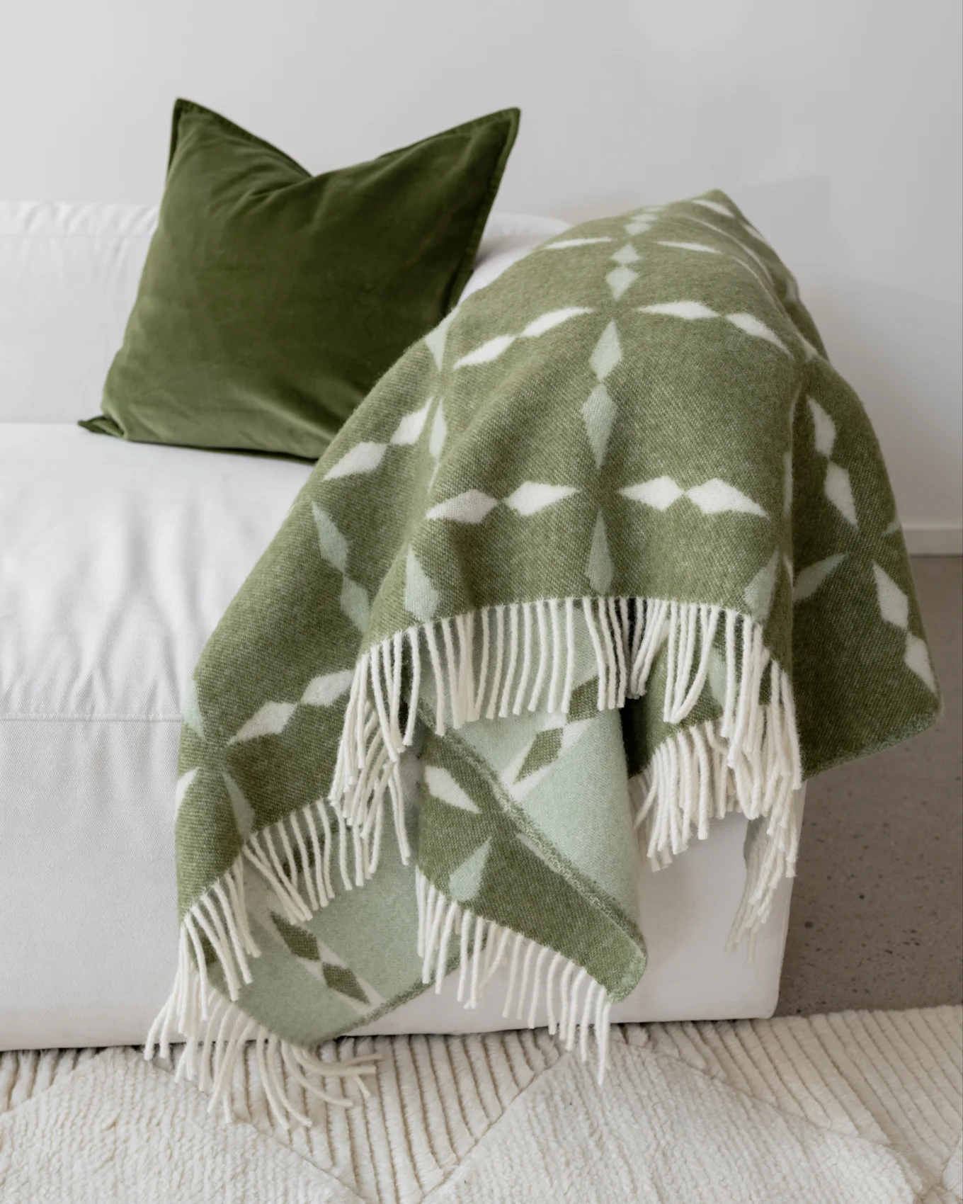 A white sofa with a dark green velvet pillow and a light green patterned throw blanket with white fringe.