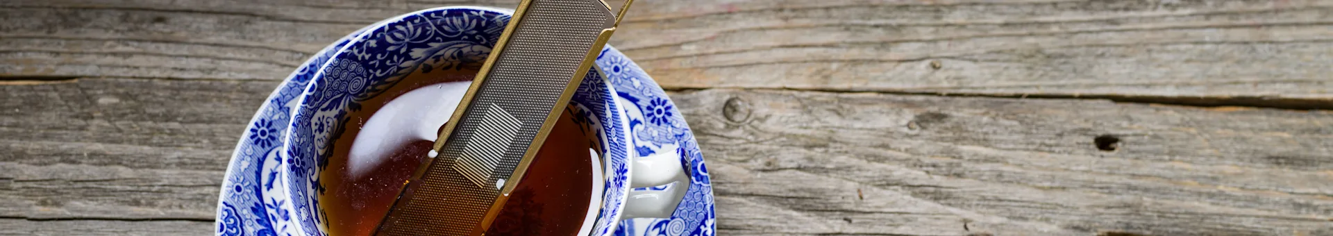 Tea infuser resting in a blue and white patterned teacup filled with tea on a wooden surface.