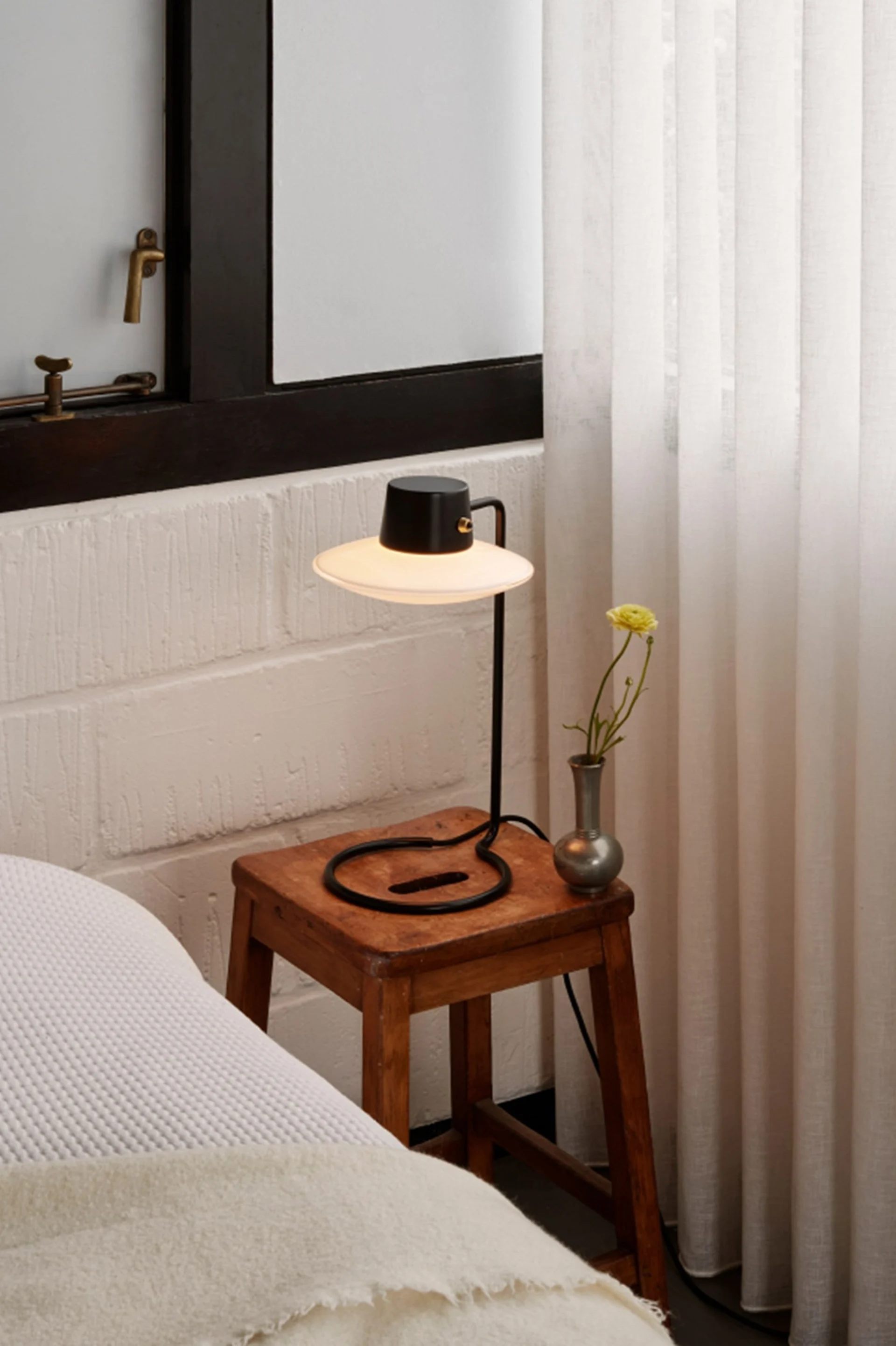 Black table lamp with a wide shade on a rustic wooden stool, a vase with a yellow flower, beside a bed and sheer curtains.