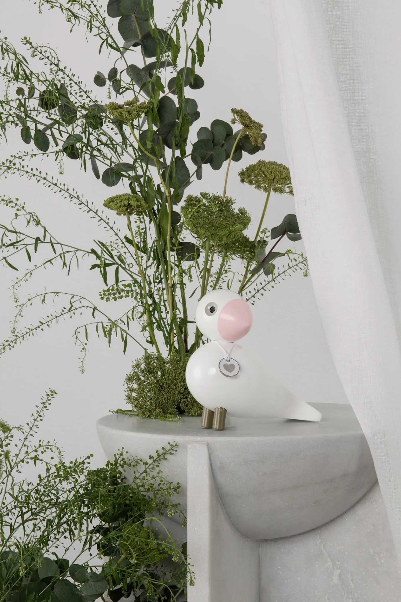 White bird figurine with a pink beak and heart charm on a grey marble stand, surrounded by green plants.