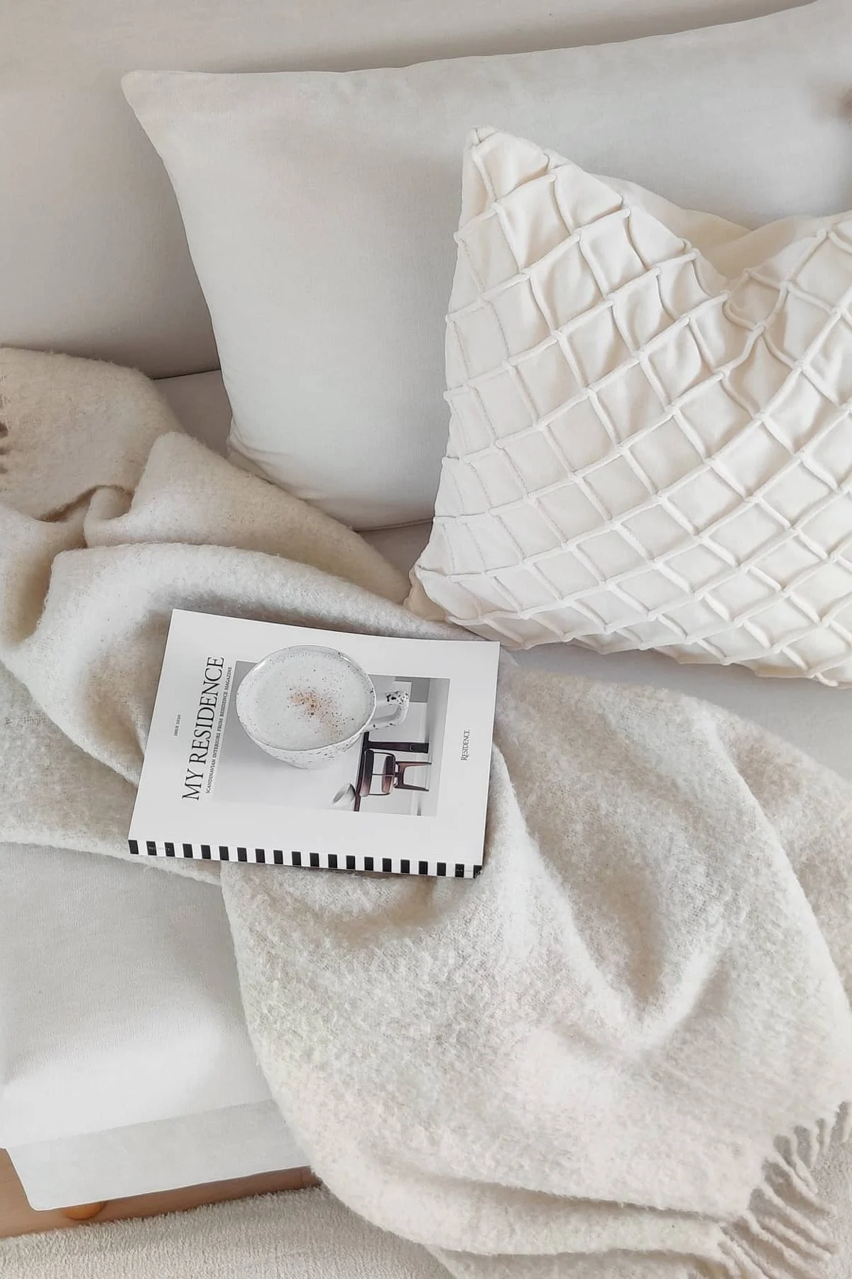 Cozy setting with a cream-colored fuzzy blanket, two cream pillows, a magazine, and a cup of coffee. One pillow has a textured grid pattern.