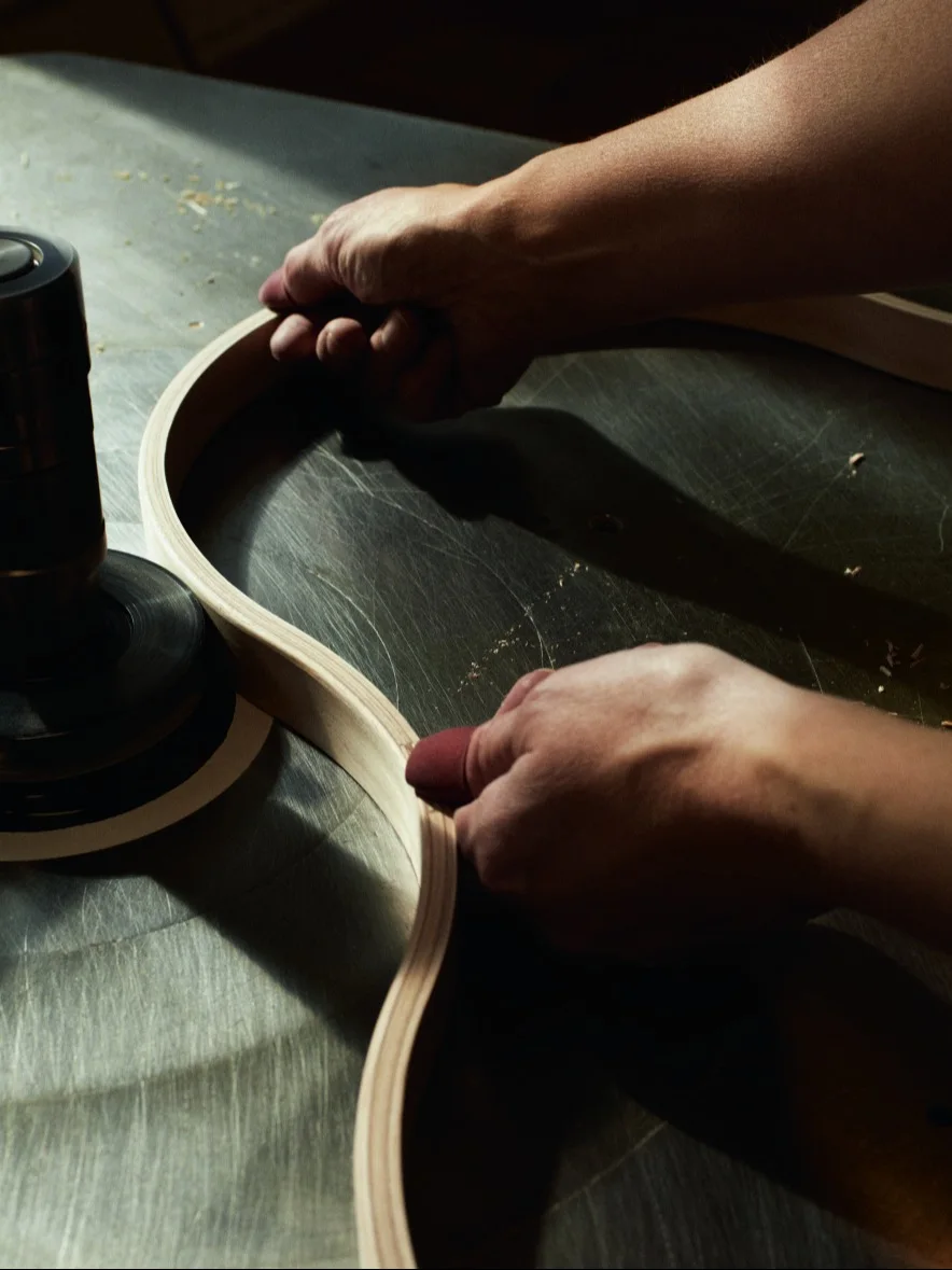 Hands sanding a curved wooden strip on a workshop machine.