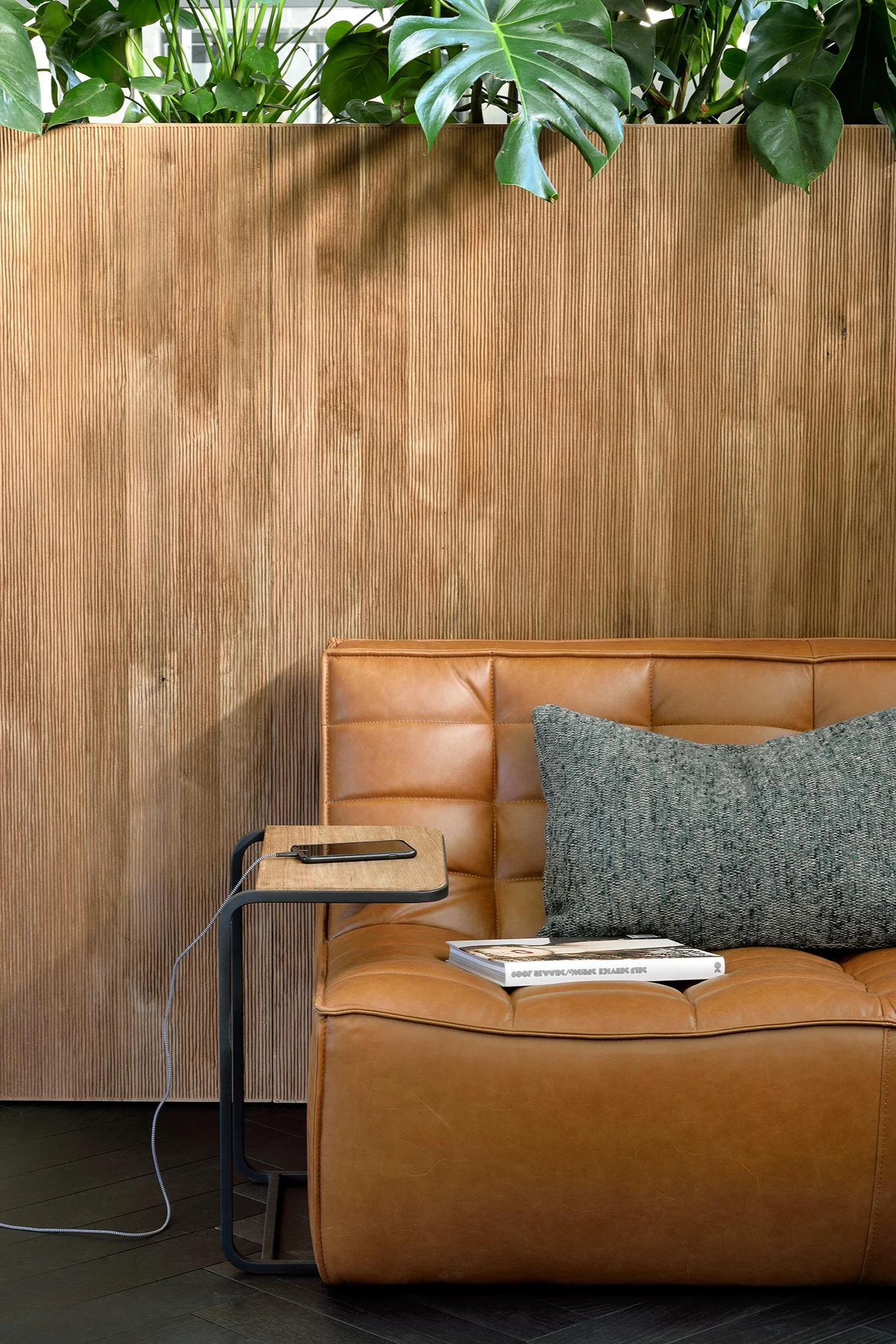 Tan tufted leather sofa with a dark grey pillow, a C-table holding a charging phone, and fluted wood wall paneling with plants above.